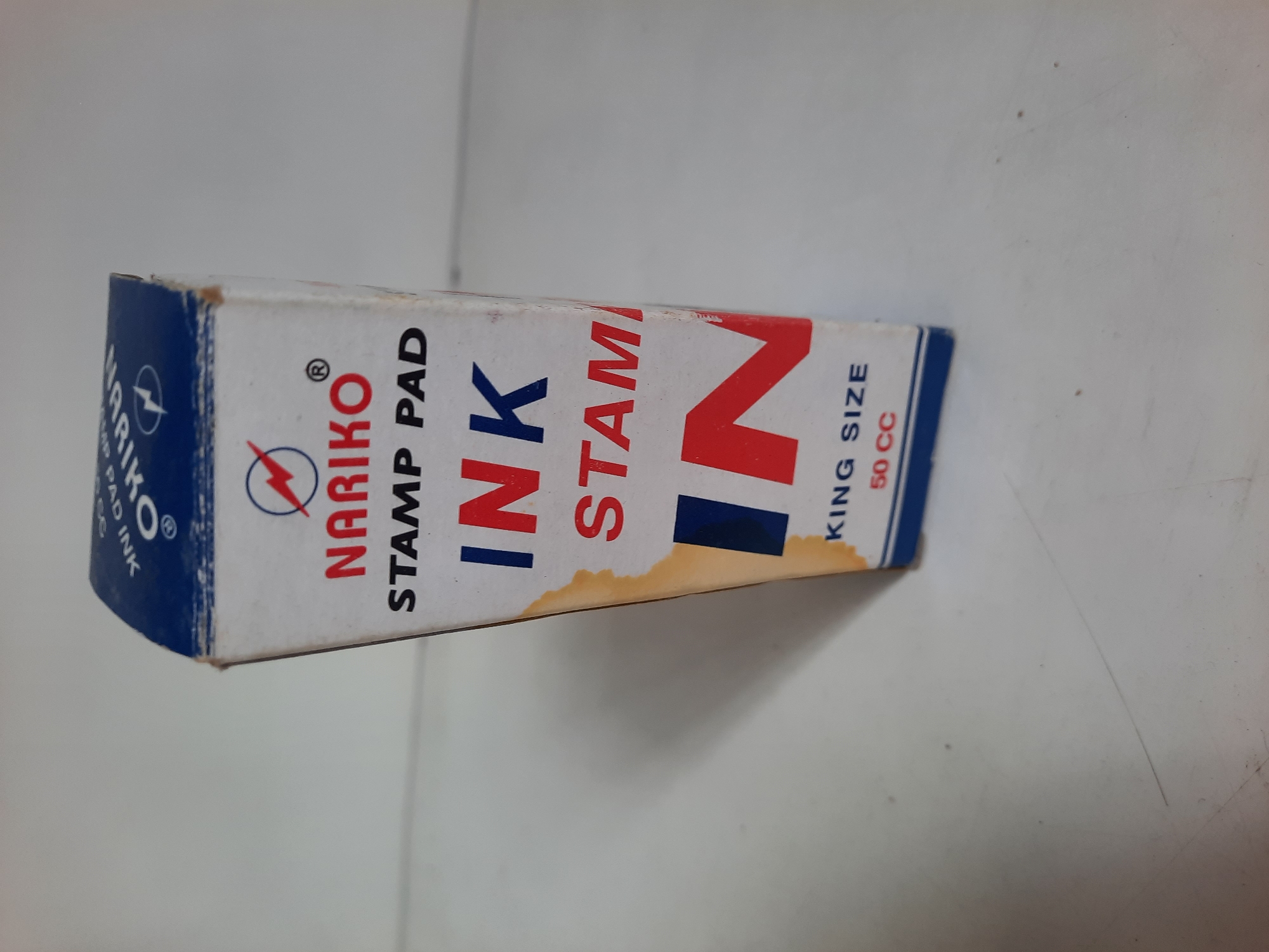 A white cardboard box for Nariko Stamp Pad Ink is placed on a white surface. The box has blue and red lettering and a red lightning bolt logo. The visible text on the box reads: "NARIKO STAMP PAD", "INK", "STAM", "IN", "KING SIZE", and "50 CC". The box is slightly worn and has a brown stain on the right side. The background is a plain white wall. The image does not provide any information about the location in Lhokseumawe, Indonesia.