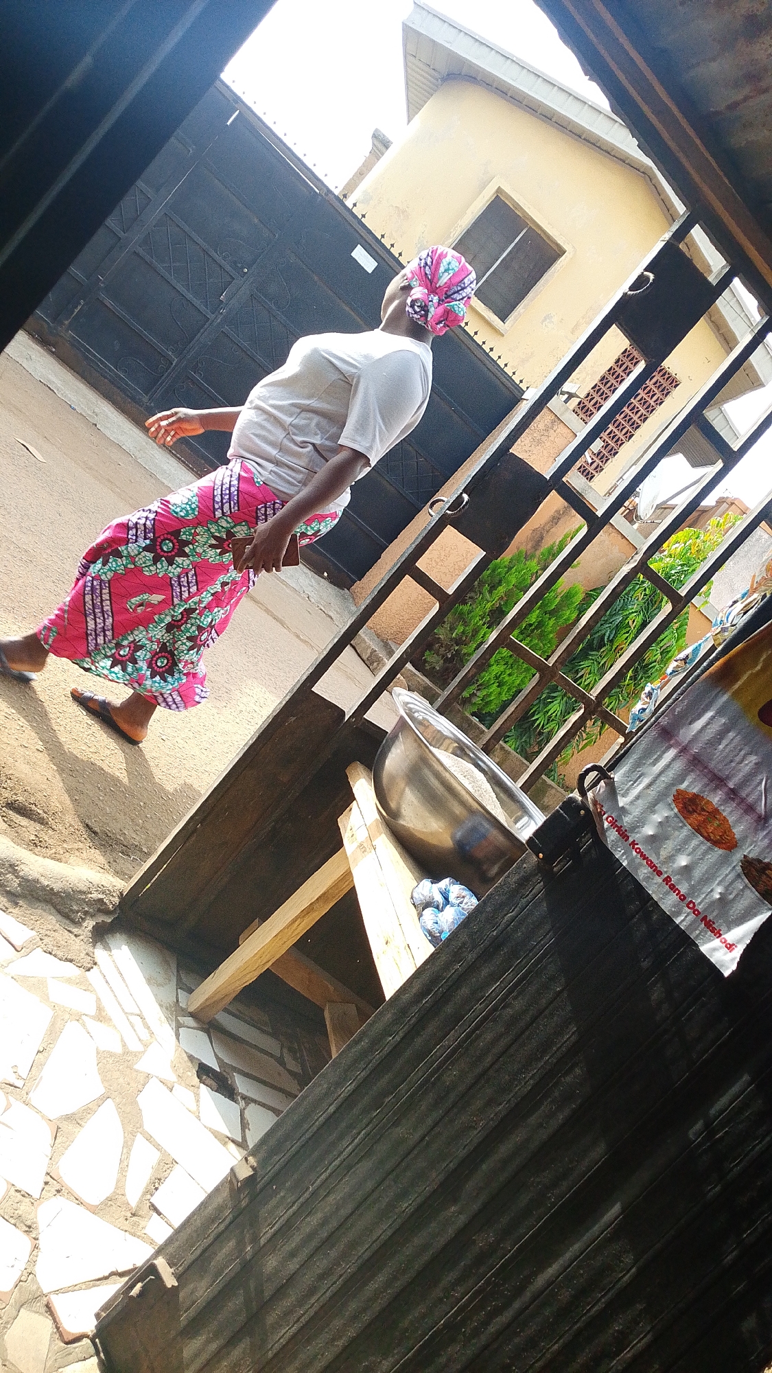 In an outdoor urban setting in an unknown city, a woman is captured in motion, her body angled away from the viewer. She wears a white t-shirt and a vibrantly patterned skirt in shades of pink, green, and blue, with a matching headscarf tied around her head. Her left arm is extended forward, and she holds a dark rectangular object, possibly a phone, in her right hand. She is walking on a paved surface adjacent to a metal fence with vertical bars and horizontal accents. Beyond the fence, a yellow building with a window and a decorative window grill is visible. Green foliage can be seen in the background. In the foreground, a metal bowl sits on wooden planks, and to its right, a white plastic bag with red lettering and images of food items is partially visible. The scene is illuminated by bright sunlight, casting shadows on the ground and creating a strong contrast between light and dark areas. The overall perspective is from a low angle, looking upwards.