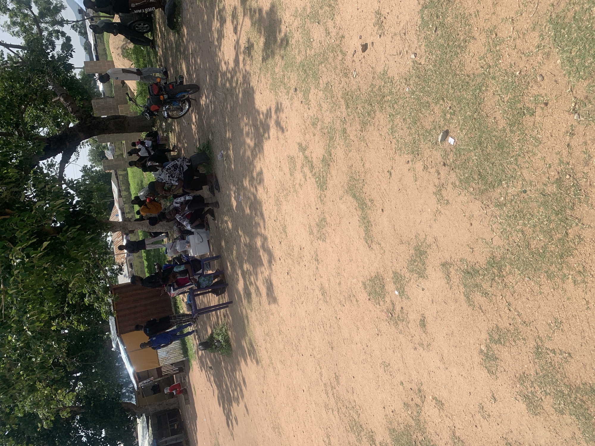 This image depicts an outdoor gathering of people in a dusty, sun-drenched environment, likely in Jalingo, Nigeria. The scene is a mix of individuals sitting and standing around what appears to be a small open area, possibly a community space or a gathering point.
Several people are seated on low benches or directly on the ground, many facing towards a central point or conversation not fully visible in the frame. Their attire suggests a casual, everyday setting. A prominent detail is a motorcycle parked near some of the individuals, indicating a mode of transportation common in the region. Behind the seated group, there are structures that look like partially built or unfinished buildings, possibly made of brick and corrugated metal, suggesting ongoing construction or a less developed area.
The lighting indicates it is daytime, with strong shadows cast by trees and structures, implying a bright, sunny day, possibly midday or early afternoon. The ground is predominantly sandy or dusty, with sparse patches of dry grass. The overall atmosphere appears relaxed and communal.
In the upper left of the image, partially obscured by a tree, is a dark vehicle with some visible text on its rear. The text appears to be "DRSTAN". The presence of the vehicle and the motorcycle, along with the simple structures and the gathered people, paints a picture of daily life in a Nigerian town. The scene is candid and natural, capturing a moment of social interaction or a community event.