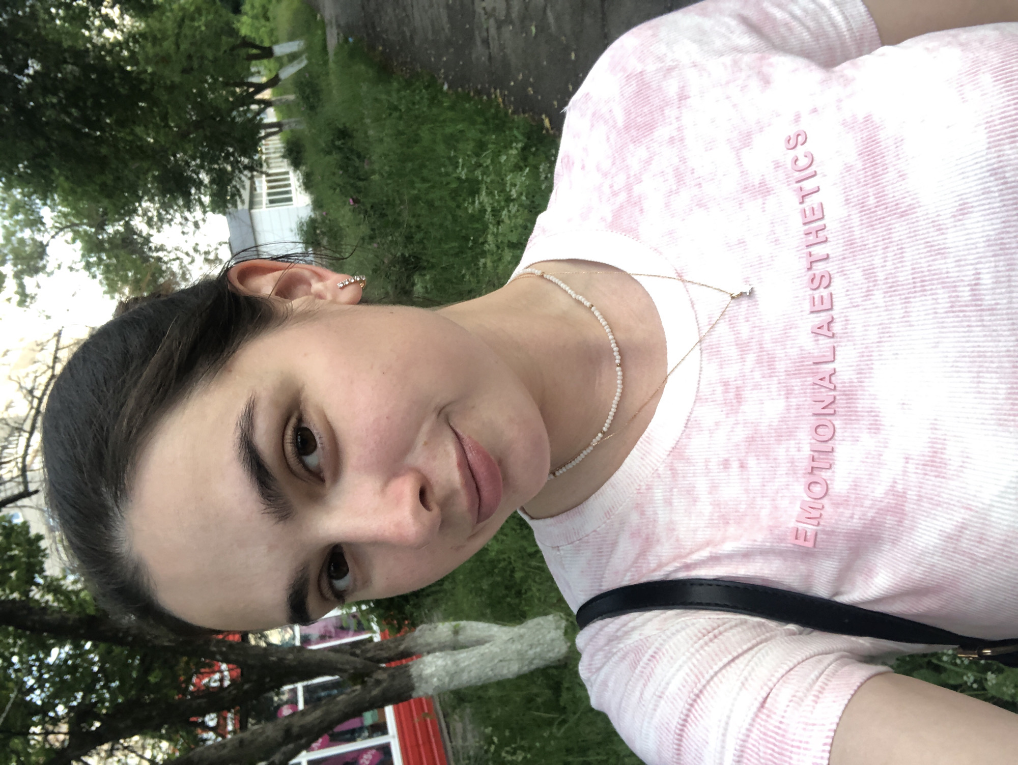 A young woman with dark hair pulled back, a fair complexion, and brown eyes is pictured from the chest up, looking slightly to her left. She is wearing a light pink tie-dye t-shirt with the words "Emotional Aesthetics" embroidered in a slightly darker pink. A thin, multi-strand beaded necklace with a small gold pendant rests on her collarbone. A black strap from a bag is visible over her left shoulder. She is standing outdoors with a blurred background of green foliage, trees, and a building with white siding and windows, typical of a residential area in Chaikovsky, Russia. The lighting suggests it is daytime, possibly late afternoon or early evening.