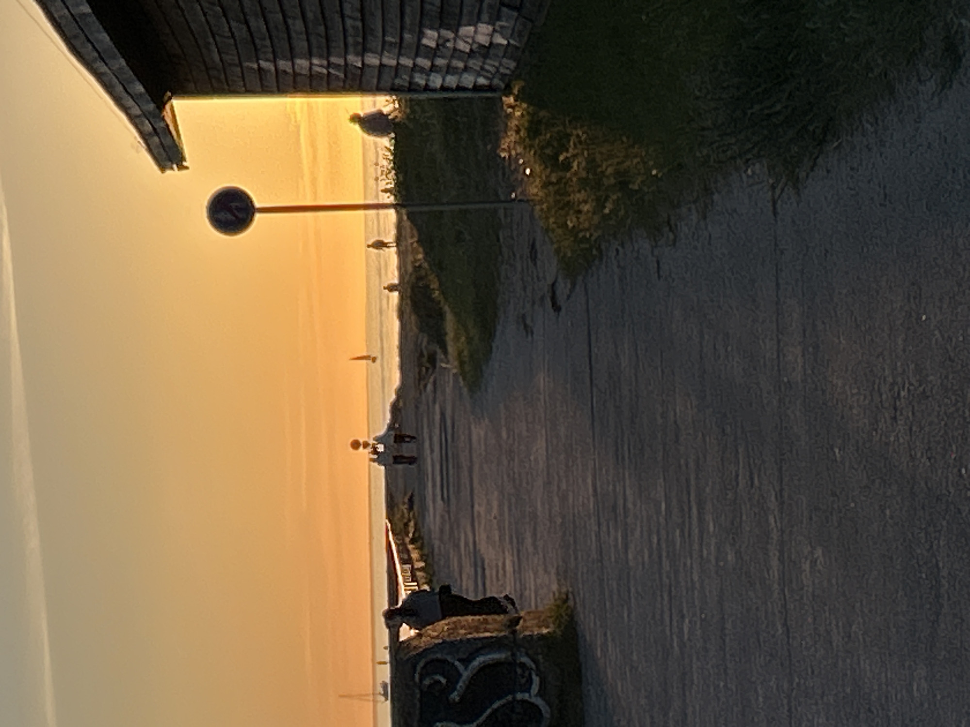 This image depicts a coastal scene during golden hour, located in Tallinn, Estonia. A wide, dark grey paved promenade occupies the lower half of the frame, extending from the foreground towards the distant horizon.
On the left side of the foreground, a dark, irregularly shaped stone structure with faded light-colored markings is visible, partially obscuring a person in dark attire who is standing beside it, facing away from the camera. Further along the promenade, two individuals are walking side-by-side, also facing away from the camera and silhouetted against the bright horizon.
To the right of the promenade, a shingled wooden building with a dark roof occupies the far right edge of the frame, illuminated by the low-angle light. In front of the building, a dark blue circular sign with a white diagonal stripe on a reddish-brown pole stands upright. Between the path and the sea, there is a grassy area with sparse vegetation. A person is seated on this grassy bank, facing the sea, and another individual stands closer to the water's edge in the middle distance.
The sea extends to the horizon, appearing calm and reflecting the warm orange-yellow hues of the sky. Several sailboats are visible as dark silhouettes on the water; one is prominent on the far left, and another is further right. The sky transitions from a bright orange-yellow near the horizon to a lighter, slightly desaturated blue-grey at the top, with a faint linear contrail visible in the upper left quadrant. The entire scene is bathed in the warm, low-angle light of either sunrise or sunset.