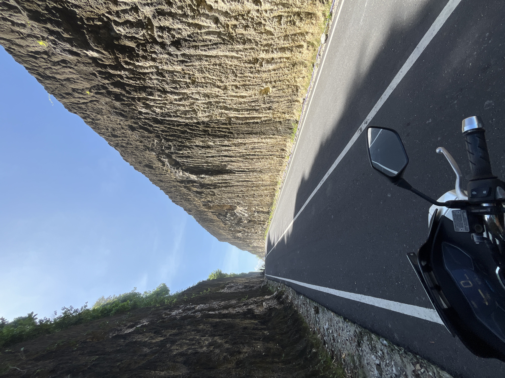 A motorcycle is parked on a paved road between high rock walls in Kuta Selatan, Indonesia. The walls are layered, and vegetation grows on the lower wall. The sky is visible at the top, with a clear blue color. The road has a painted white line separating lanes.