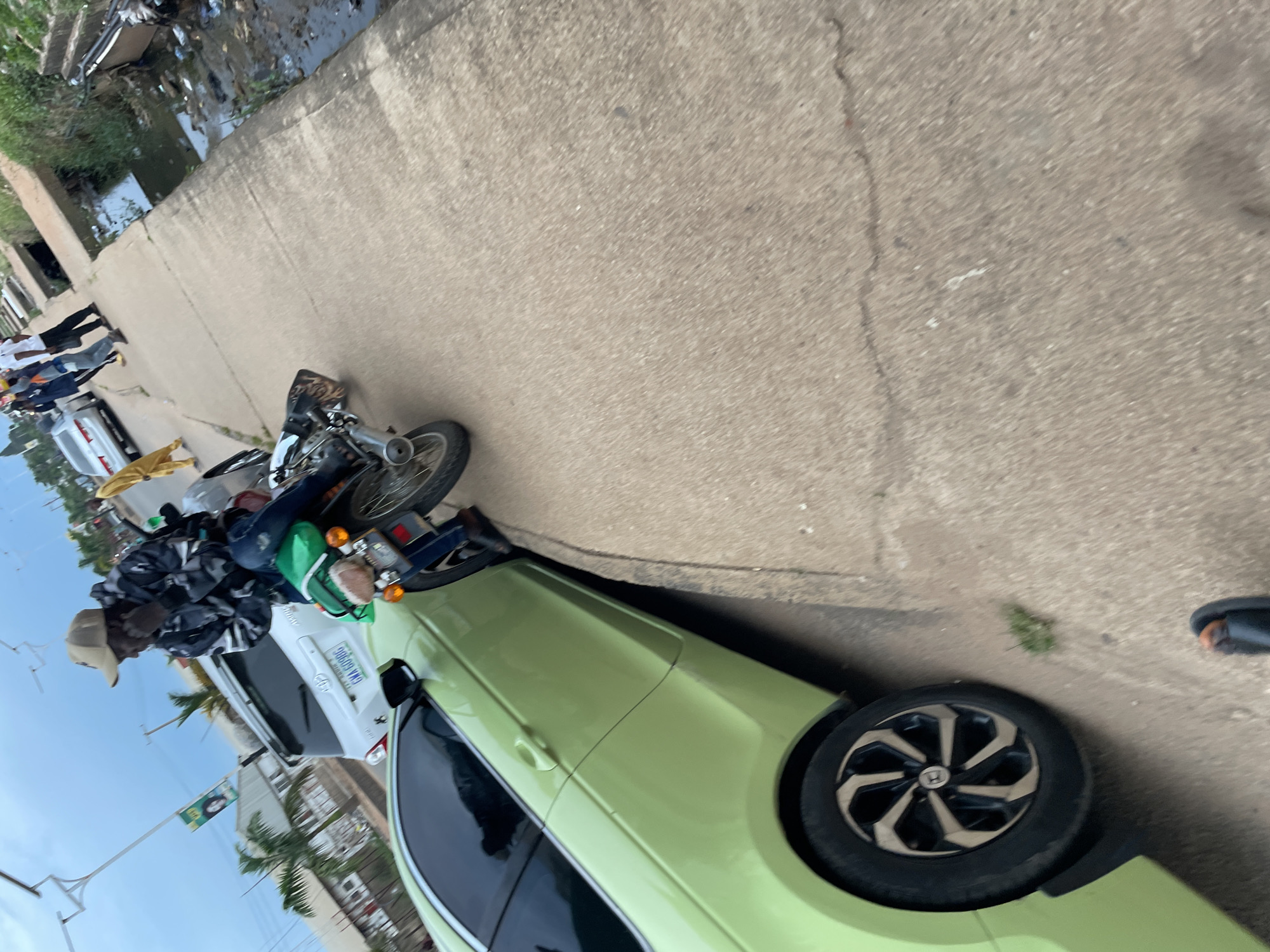 In Barakin Lerawi, Nigeria, a person wearing a camouflage jacket and a beige cap is seated on a green motorcycle. The motorcycle is parked alongside a pale green car. In the background, several other vehicles and pedestrians are visible, suggesting a street scene. The environment includes a paved walkway adjacent to a drainage ditch filled with murky water and debris. The sky is overcast.