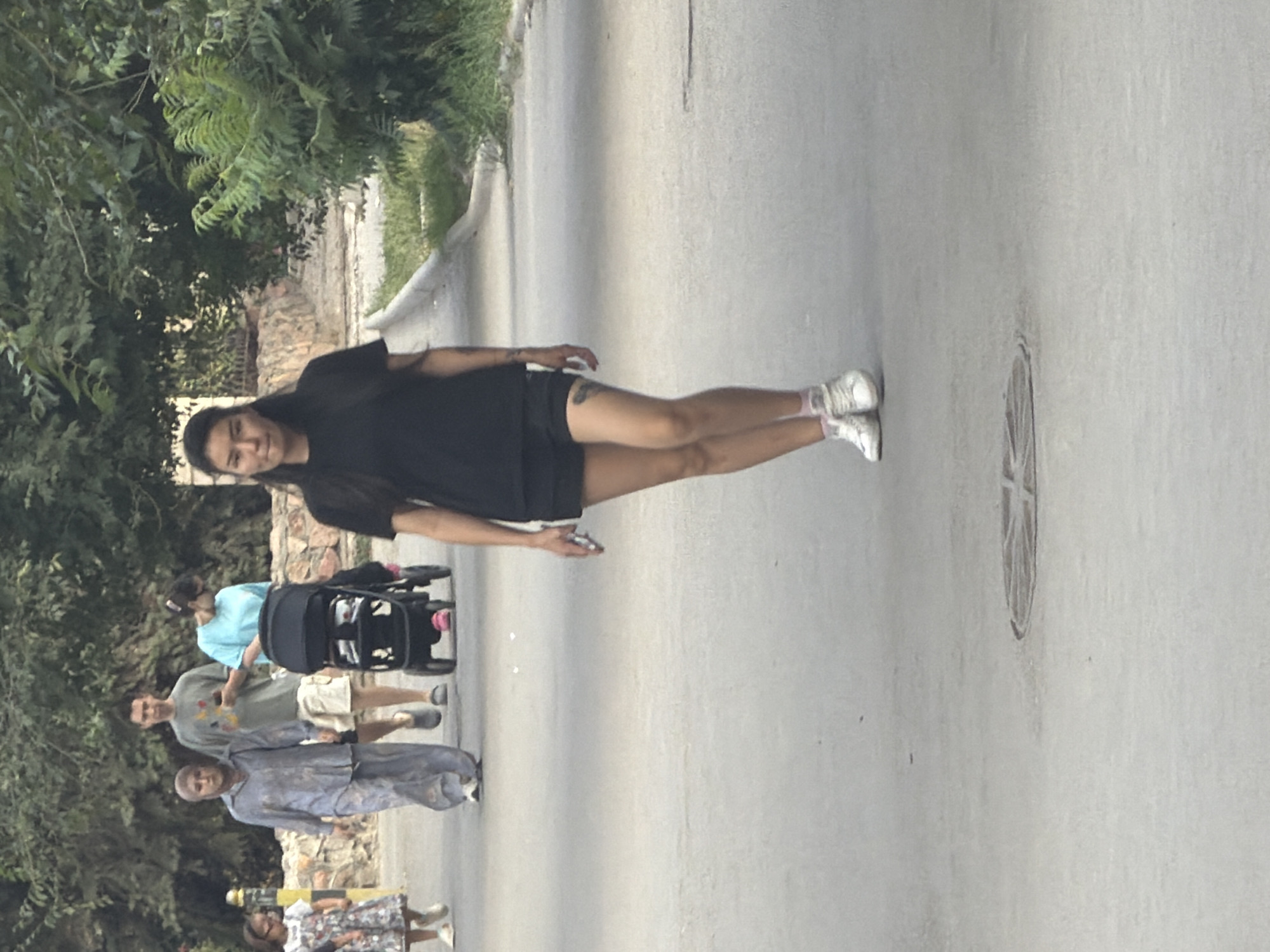 An outdoor full shot shows several people walking on a paved surface, possibly a sidewalk or park path. In the foreground, a woman with long dark hair is walking towards the camera, wearing a black t-shirt and black shorts with white sneakers and pink socks. She has tattoos on her left leg and is holding a mobile phone in her right hand. Behind her, a family walks away from the camera. A woman in a light blue t-shirt is pushing a black stroller with pink accents. Next to her, a man in a green t-shirt and khaki shorts is holding his phone. To their left, a woman in a grey abaya and hijab walks with her head down. On the far left, a young girl in a patterned dress and white shoes is walking ahead of the group. The background is filled with lush green trees and foliage, with a stone wall visible behind the family. The ground in the foreground has a circular metal manhole cover.