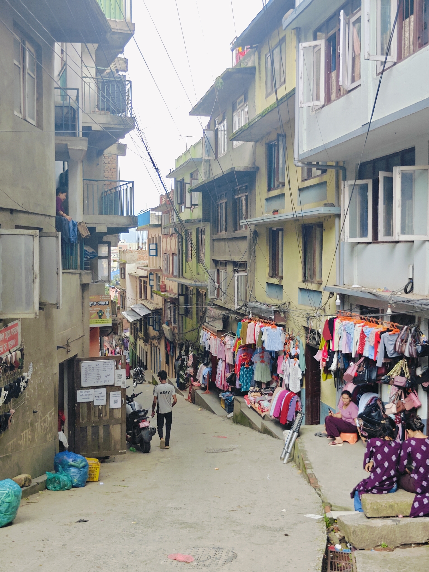 The image depicts a narrow street in Kirtipur, Nepal, lined with multi-story buildings. The buildings exhibit a variety of architectural styles and colors, with some featuring balconies and open windows. Clothes and bags are displayed for sale on racks along the side of the street, suggesting a market area. Several people are present: a man wearing an "ARMY" t-shirt walks away from the camera, a woman sits on a small stool looking at her phone, and two other individuals in purple outfits are seated on concrete blocks. A motorcycle is parked near a wooden door. Trash bags and a yellow crate are visible on the left side of the street. Overhead, a complex network of electrical wires crisscrosses the sky. The overall scene suggests a bustling urban environment with commercial activity and residential spaces integrated.