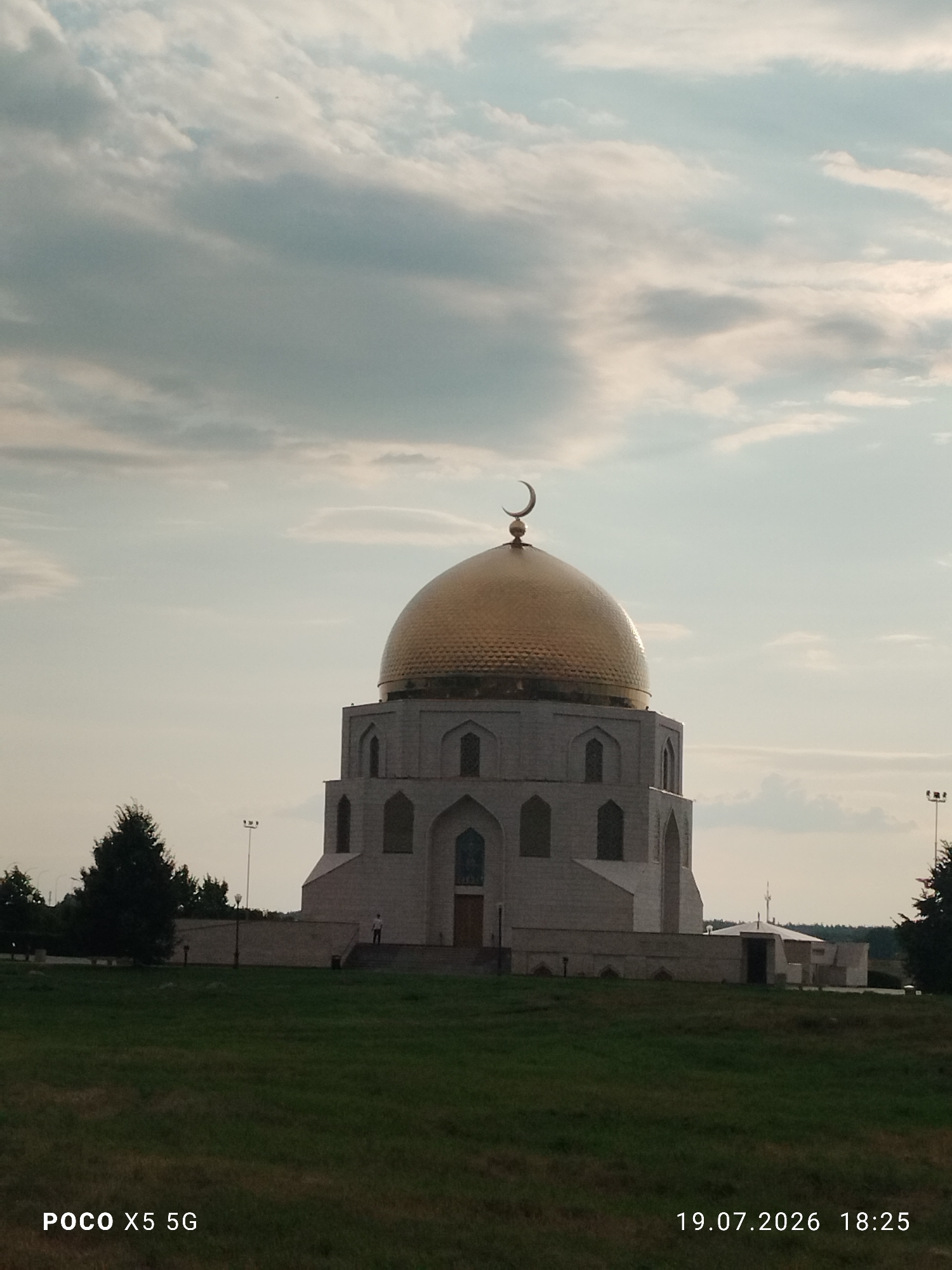 The image shows a large, ornate building, likely a mosque, with a golden dome topped with a crescent moon. The building is constructed of light-colored stone and features arched windows. It is situated in a grassy, park-like area with trees in the background. The sky is partly cloudy. In the lower left corner, text reads "POCO X5 5G", and in the lower right corner, text reads "19.07.2026 18:25". The building appears to be the Memorial Mosque of the Volga Bulgars in Bolgar, Russia, a UNESCO World Heritage site. A single person is visible standing on the steps leading to the entrance of the building.