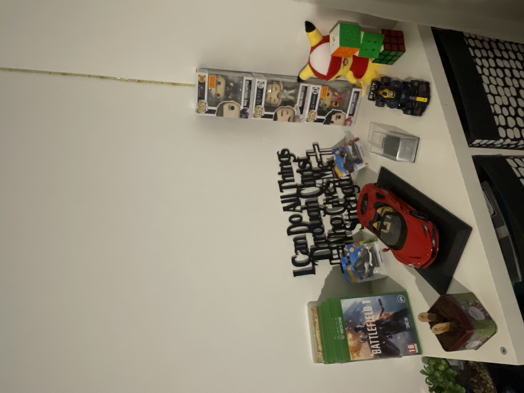 The image depicts a shelf against a light-colored wall. On the shelf are several items, including Funko Pop figures, a Pikachu plush with stacked Rubik's Cubes, a toy Formula 1 car, a quote sign, die-cast cars, a red toy car on a stand, a copy of the video game "Battlefield 1" for Xbox One, and a decorative pot with a plant.