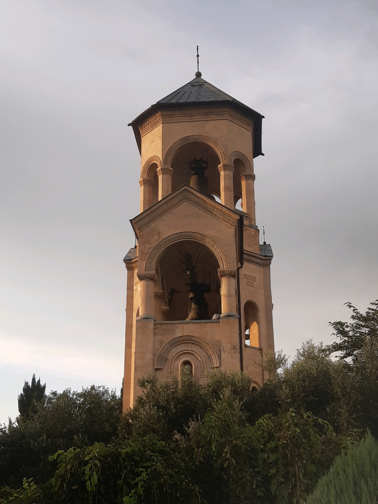 The image shows the bell tower of the Holy Trinity Cathedral of Tbilisi, also known as Sameba Cathedral, in Tbilisi, Georgia. The bell tower is a tall, multi-tiered structure made of light-colored stone. It features arched openings at various levels, and a dark, conical roof topped with a spire. Bells are visible within the arched openings. The tower is surrounded by lush green trees and foliage, with a cloudy sky in the background. The lighting suggests it might be late afternoon or early evening, with the warm tones of the stone indicating the sun is low on the horizon.
