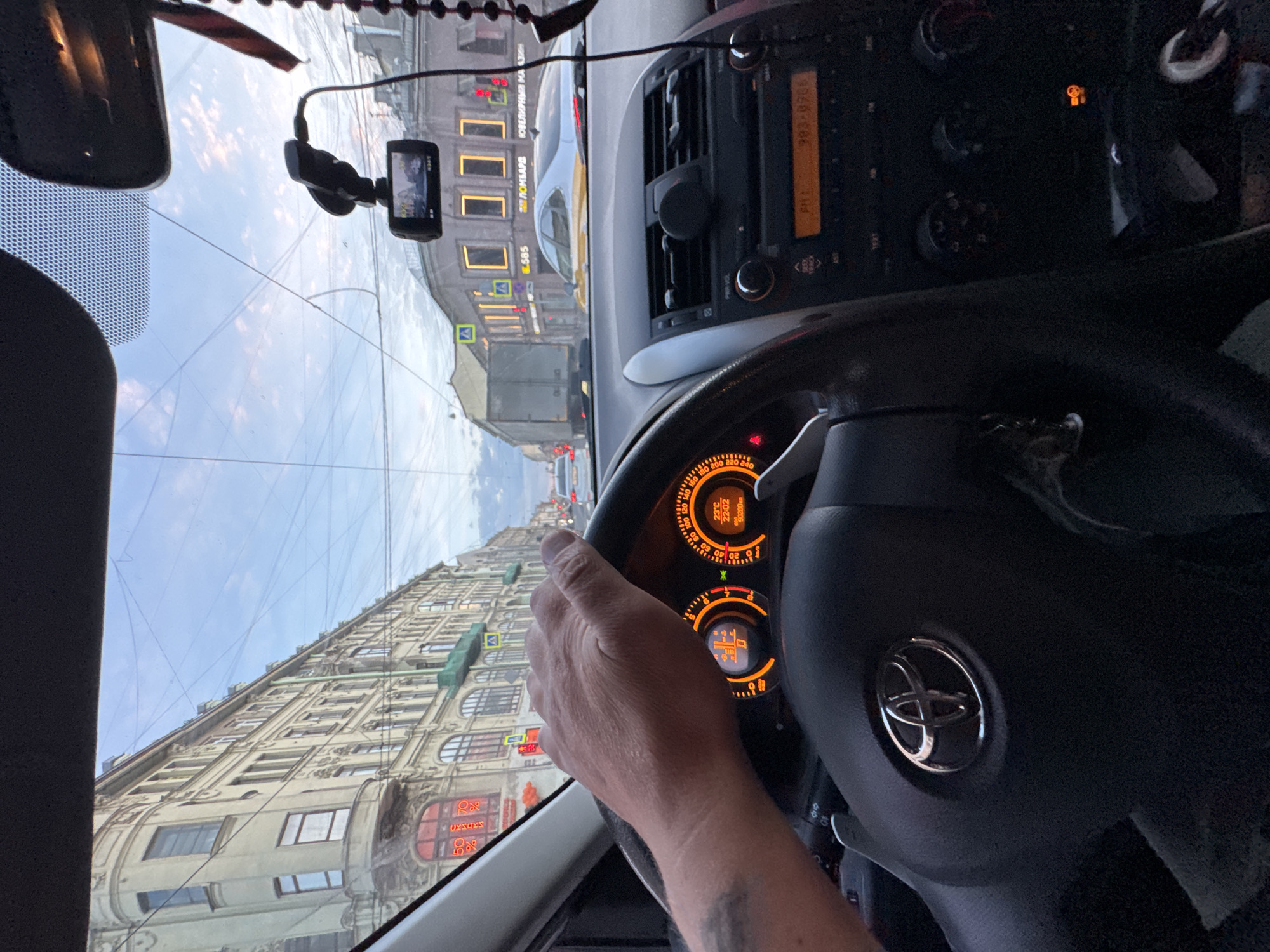 The image provides a driver's perspective from inside a car on a city street in Saint Petersburg, Russia. The sky, seen through the windshield, is partially cloudy, suggesting either early morning or late evening. The car's illuminated dashboard clock confirms a late hour, showing "22:02".
In the foreground, a person's right hand rests on the steering wheel, which bears a prominent silver Toyota logo. The dashboard features an illuminated, orange-themed instrument cluster displaying a speedometer, various warning lights, and digital readouts including an odometer (showing "90280") and an exterior temperature of "22°C". To the right, the car's audio system or infotainment unit is visible, displaying "FM1" and the frequency "903-0708". A dashboard camera is mounted on the windshield, capturing the road ahead.
Beyond the windshield, the scene reveals a classic urban setting. Multi-story, ornate buildings with numerous windows line the street, their architectural style typical of historic European cities. Overhead power lines, commonly found for public transport like trams or trolleybuses in Russian cities, crisscross the sky. Visible text on the buildings includes "ПЛОМБАРД" (Pawnshop) and "ЮВЕЛИРНЫЙ МАГАЗИН" (Jewelry Store) in Cyrillic, firmly placing the scene in Russia. Road signs, including pedestrian crossing indicators and a yellow diamond sign for a main road, are also discernible. The overall impression is one of being stopped or slowly moving within an active urban environment.