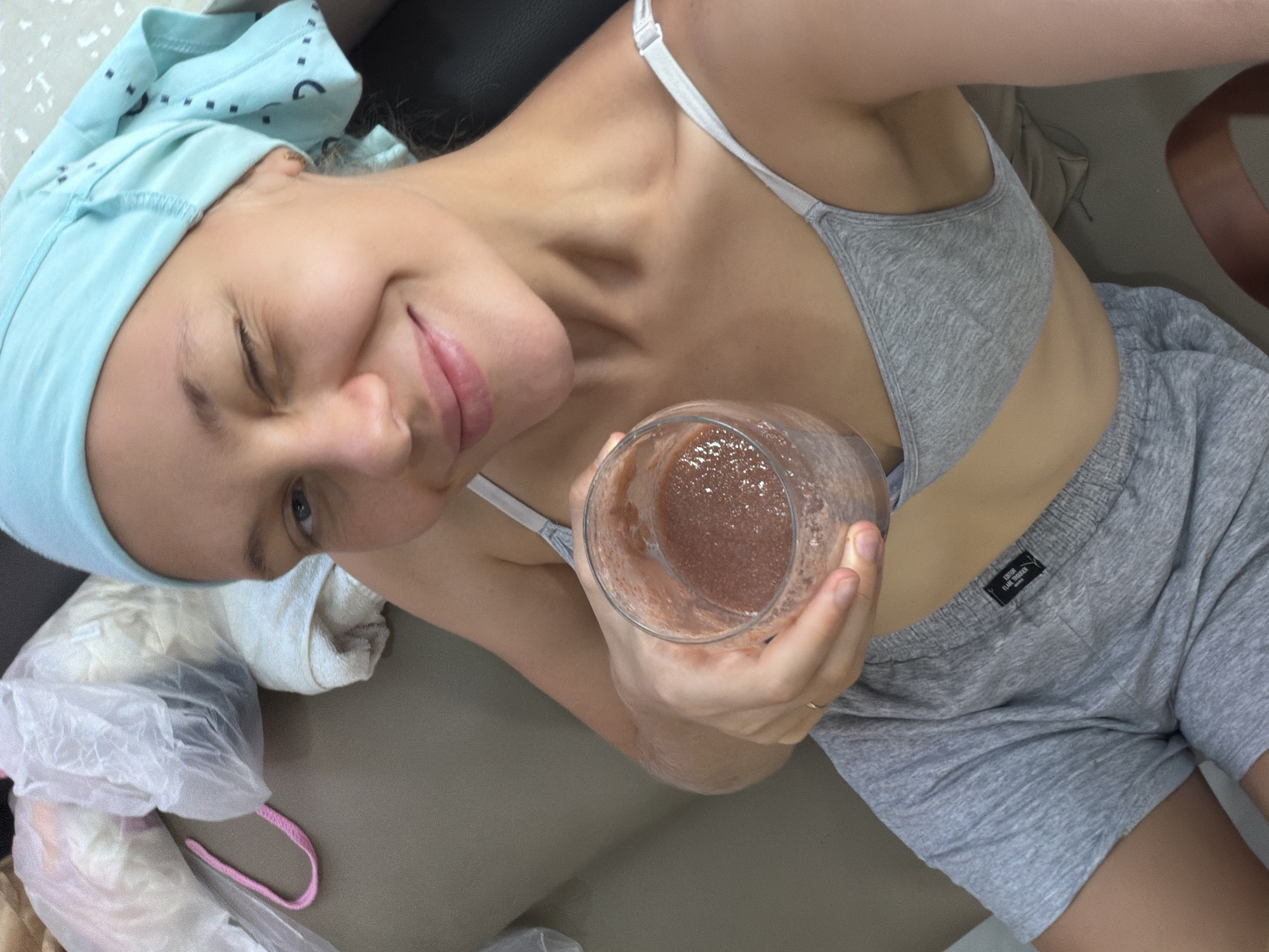 The image shows a young woman with a light blue headscarf and a grey sports bra and shorts. She is holding a glass of a pinkish smoothie in her right hand and winking at the camera. The background is out of focus, but appears to be an indoor setting with some plastic bags and white fabric. There is no indication of the location being Bac Nha Trang Ward, Vietnam.