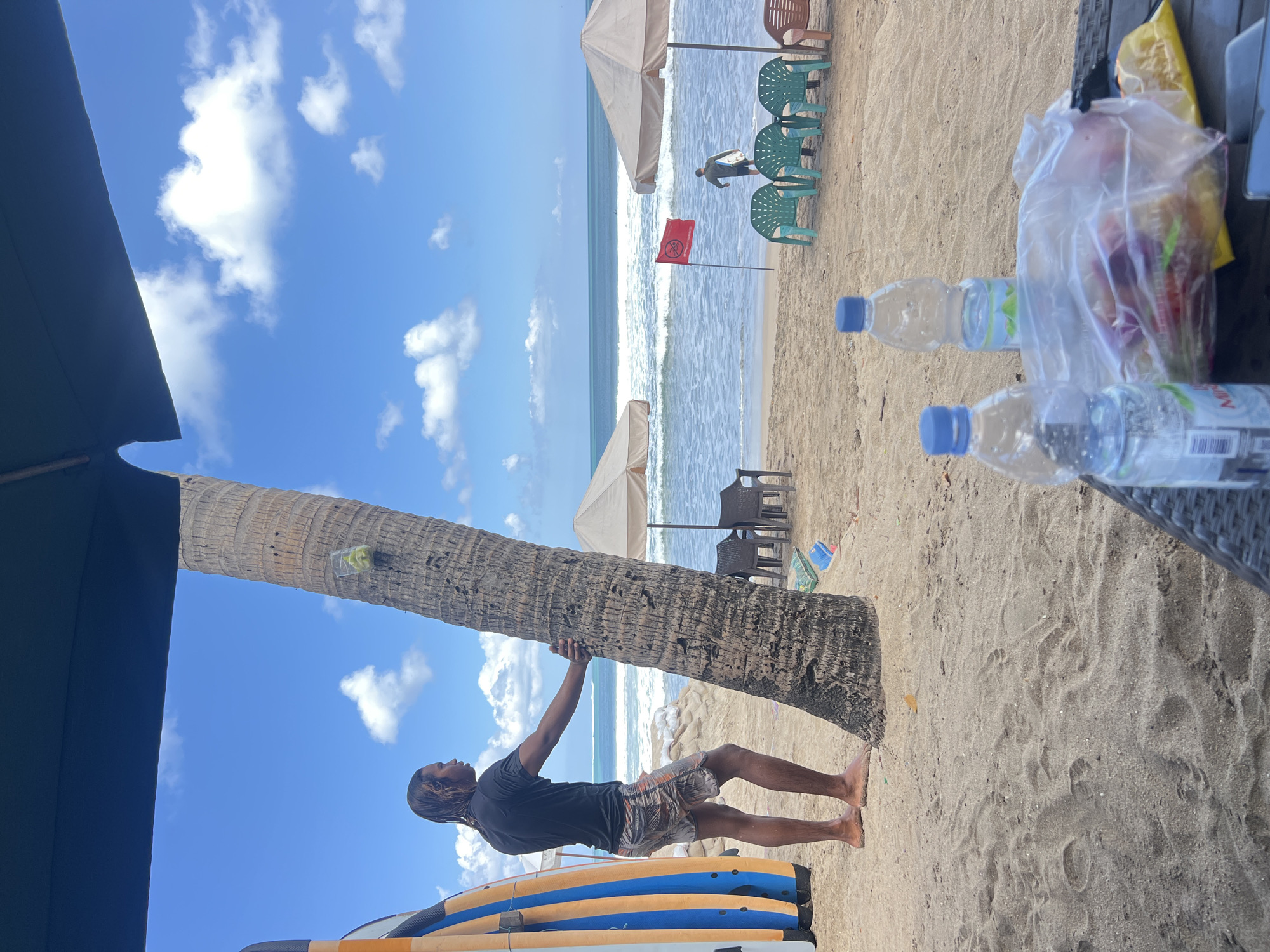 A man with long, dark, wet hair stands next to a palm tree on a sandy beach in Kuta, Indonesia. He is wearing a black t-shirt and patterned shorts. Two surfboards, one orange and blue, the other orange and white, lean against the left side of the frame. In the background, the ocean with moderate waves meets a bright blue sky with scattered white clouds. Two beige beach umbrellas are set up on the sand, with chairs underneath them. A red flag with a white symbol is planted in the sand near the water's edge. Another person, holding a surfboard, walks towards the ocean. In the foreground, two plastic water bottles and a clear plastic bag filled with fruit are on a table. The scene is bathed in daylight, suggesting it is a sunny day.