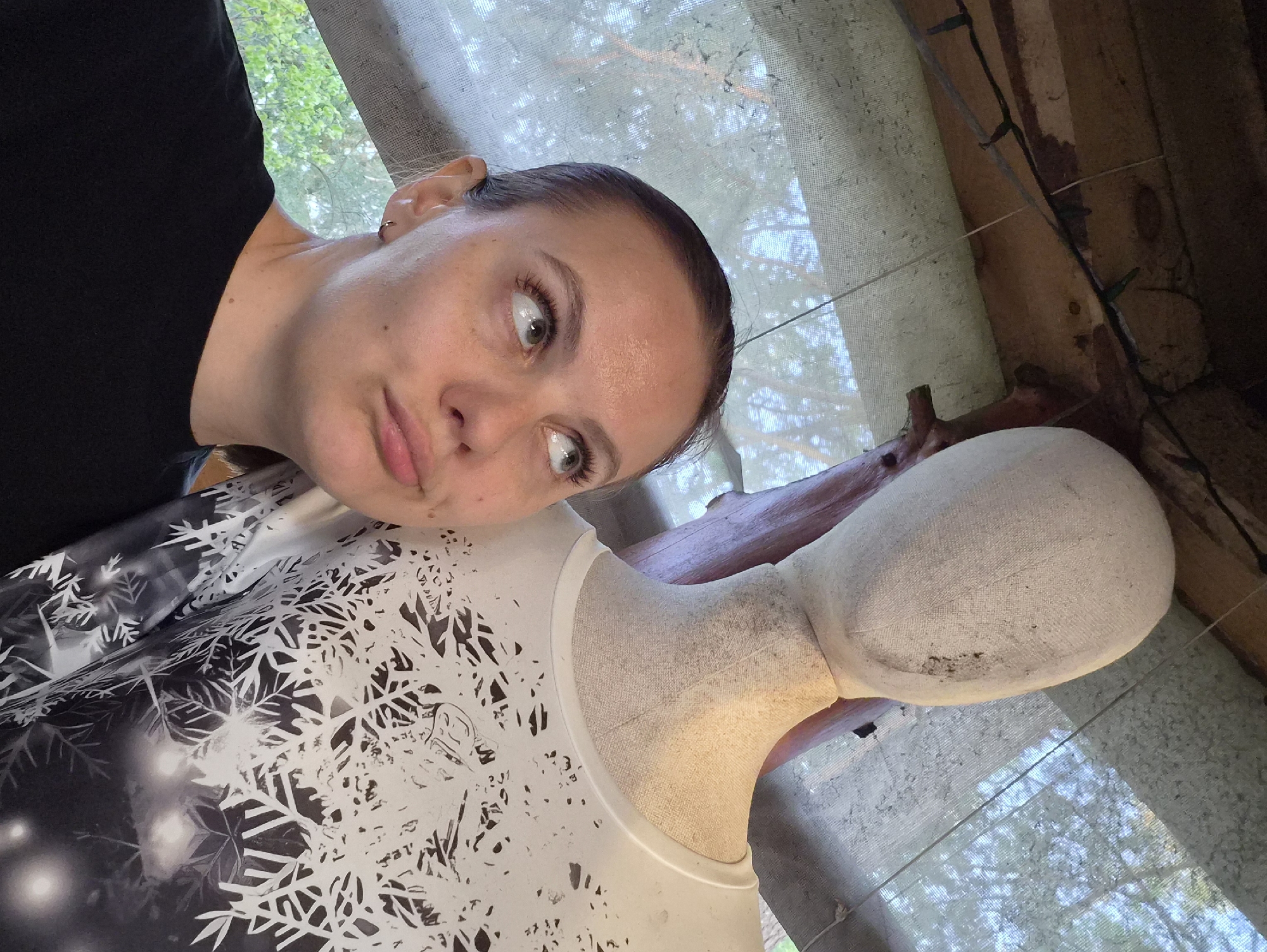 A woman in a black shirt is leaning towards a dress form wearing a white dress with black and white snowflake patterns. The woman has light skin, brown hair pulled back, and light green eyes. She has a thoughtful expression. The dress form is positioned in front of a window covered by a light-colored curtain. The backdrop includes the interior of what appears to be a wooden structure, with exposed beams and what may be string lights visible. There is no verifiable information to suggest that the location is within the city of Прикамский, Russia.