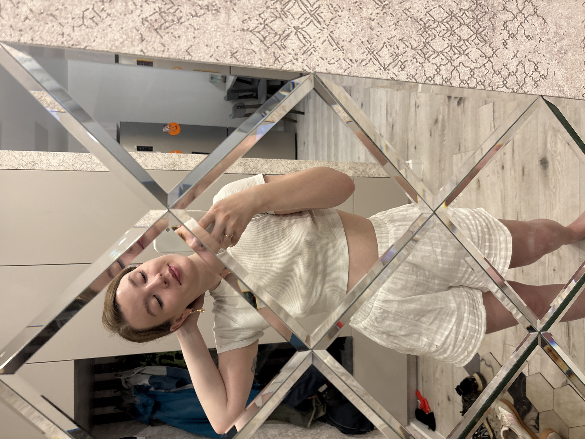 A person wearing white shorts and a white top is reflected in a diamond-patterned mirror. The mirror's reflection also shows a room with a closet underneath a white surface, possibly a countertop. The floor appears to be a light-colored wood or tile. The location, Izhevsk, Russia, cannot be confirmed from the visual information.