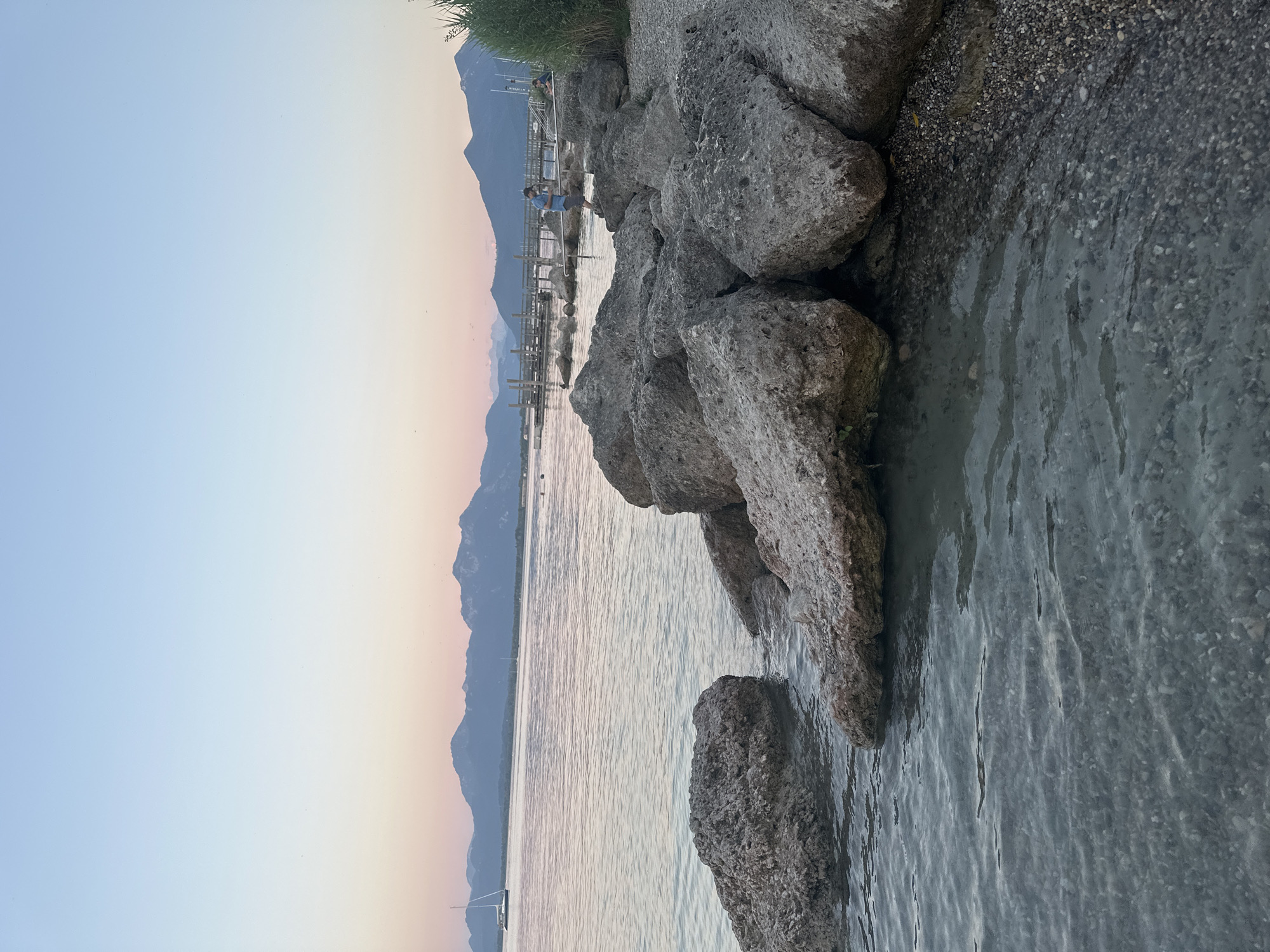 The image depicts a tranquil lakeside scene at dusk, likely in Prien am Chiemsee, Germany. The sky is a gradient of soft pinks and blues, with distant mountains silhouetted against the horizon. A calm body of water, possibly Lake Chiemsee, occupies the majority of the frame, reflecting the subtle colors of the sky. Large, rough-textured rocks are scattered along the shoreline, with smaller pebbles and gravel visible where the water meets the land.
In the middle ground, a wooden pier extends out into the lake. A person wearing a blue shirt and shorts is standing on the pier, facing away from the camera, seemingly engaged with something on the pier or in the water. Another person, partially obscured by vegetation, sits on the pier to the right. A sailboat with its mast visible is anchored in the water to the left, and another small boat can be seen further out. Lush green vegetation lines the far right edge of the image. The water is clear enough to see the gravelly bottom near the shore.