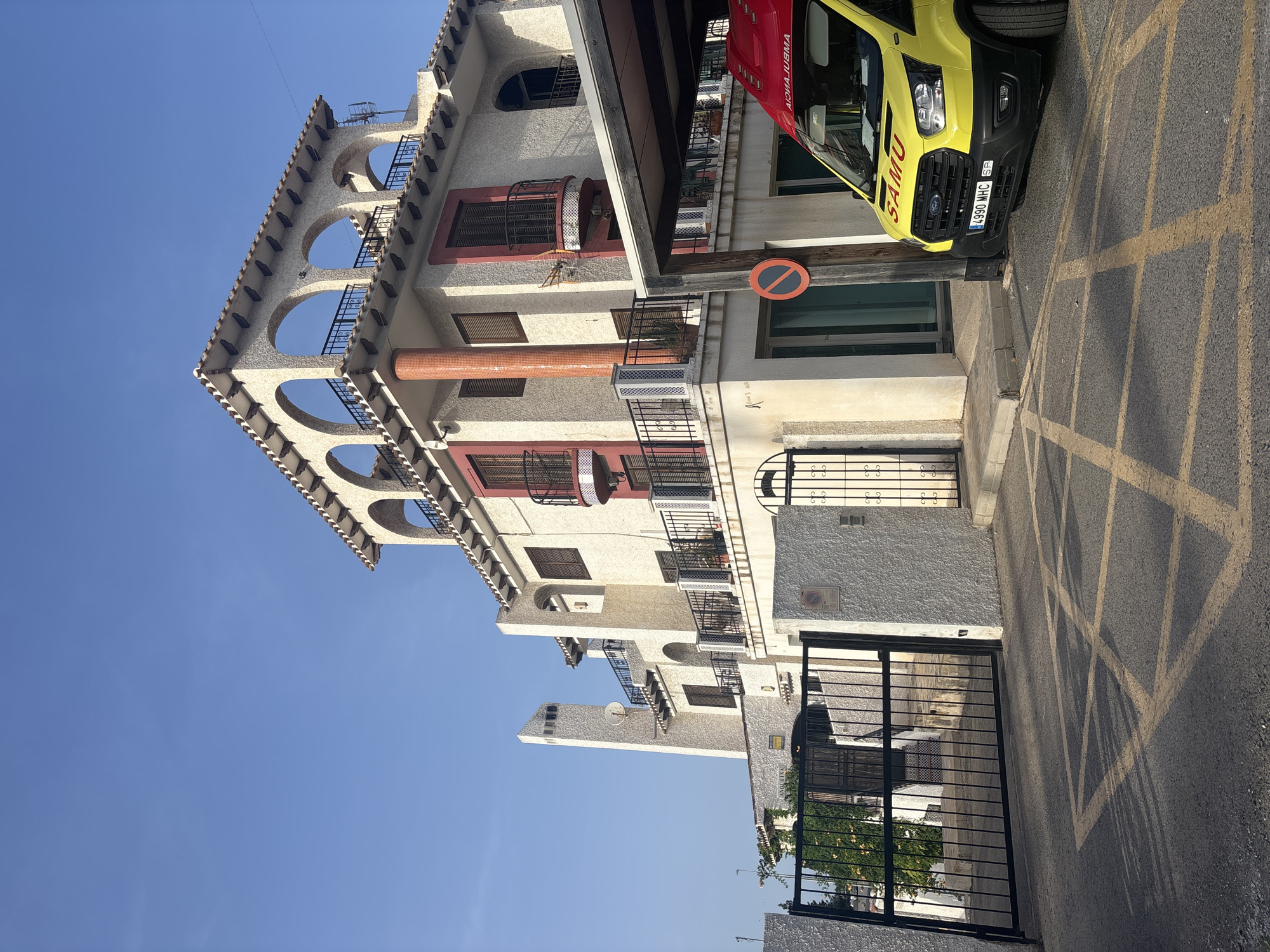 The image depicts an exterior view of a building in Orihuela, Spain, with an ambulance parked in front. The building is multi-story, featuring a white stucco facade with red accents around windows and balconies. The architecture includes arches on the top floor, a distinctive chimney, and wrought-iron railings. A black metal gate is visible to the left, leading to a pathway. In the foreground, a yellow ambulance with "SAMU" written on it is parked to the right, under a covered area. The ground in front of the building and ambulance is asphalt with yellow painted lines, possibly indicating a parking area or no-parking zone. The sky is clear and blue, suggesting daylight.