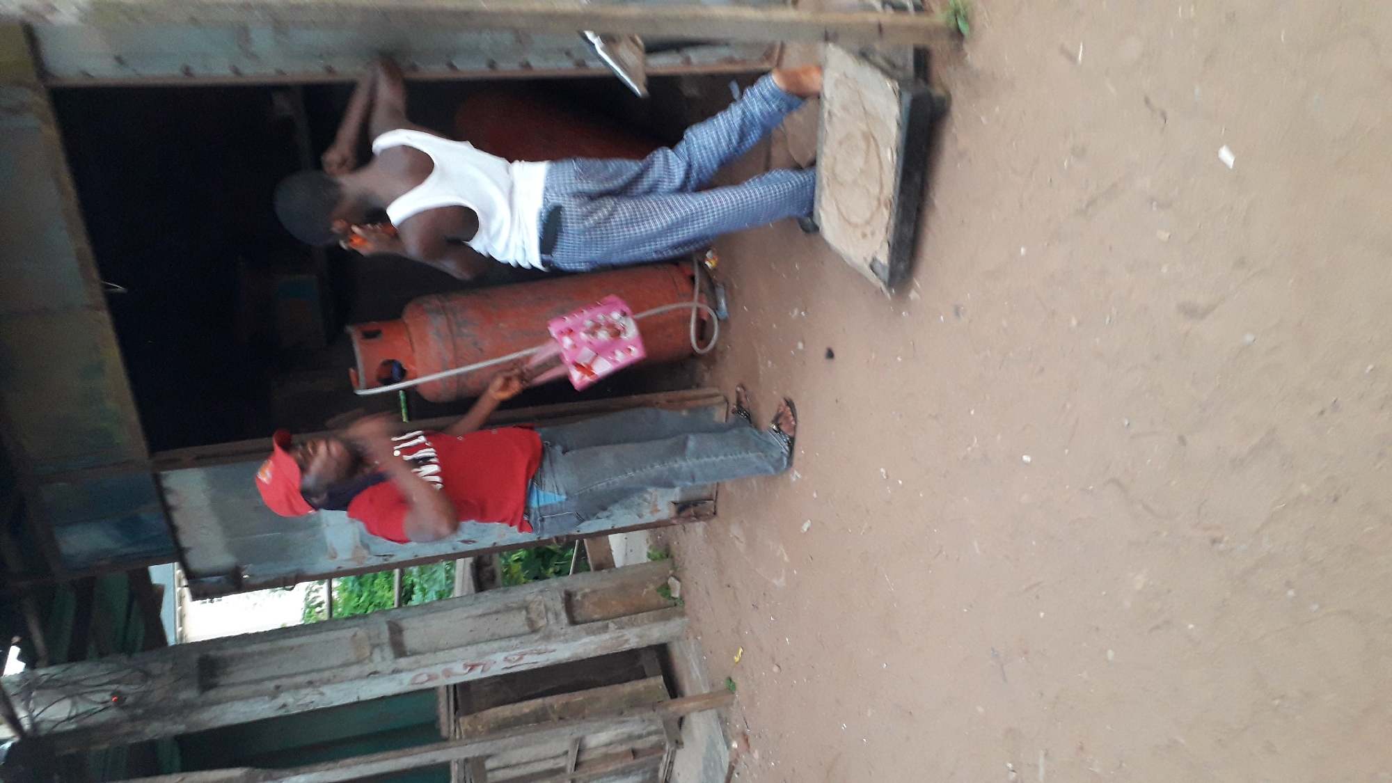 The image shows two men outside of a building. One man is wearing a white tank top and plaid pants, while the other is wearing a red shirt and blue jeans. There is a gas tank next to them, and one man is holding a pink purse. The ground appears to be dirt. The location is in Nkpor, Nigeria.