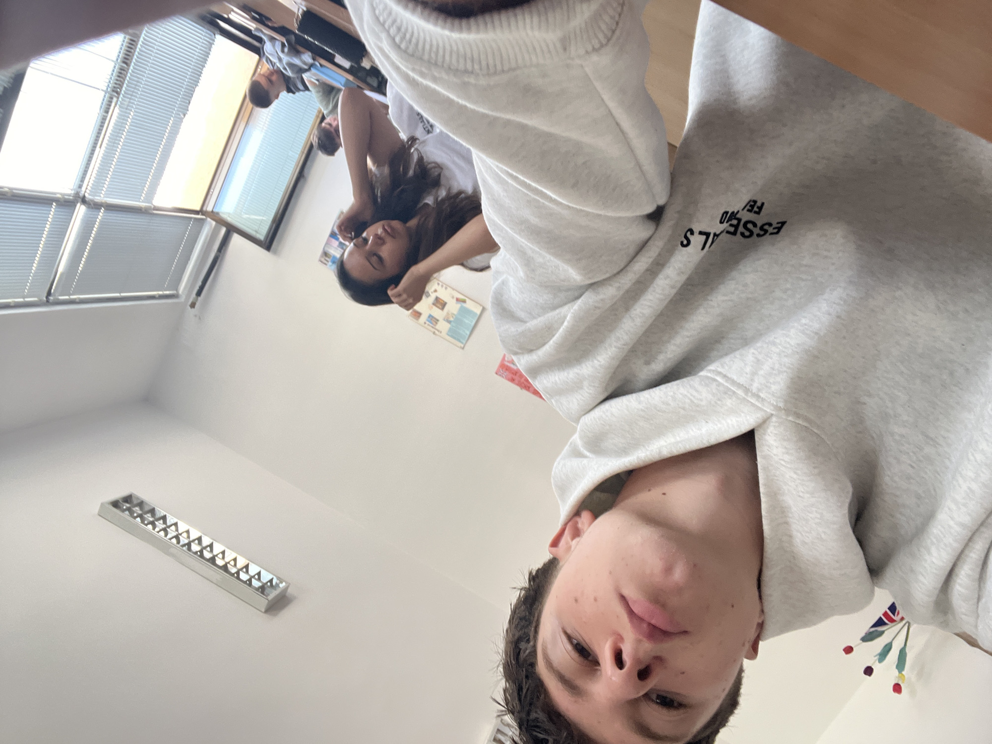 This media file depicts a casual scene within what appears to be a classroom in Zlatograd, Bulgaria, captured from a low-angle perspective. The image is oriented sideways, but if rotated, it shows a young person in the foreground taking a selfie.
The primary subject, likely a young male, is in the immediate foreground, looking directly at the camera with a neutral to slightly playful expression. He has short dark hair and is wearing a light grey hoodie. Text printed on the hoodie's sleeve is visible, reading "ESSENTIALS FE/F" (or similar, due to the original orientation).
In the mid-ground, another young person, a female with long dark hair, is visible, wearing a white t-shirt. She appears relaxed, possibly resting her head on her arm or a desk, and is looking downwards or to the side. Further in the background, the head and shoulder of a third person can be partially seen.
The setting is an indoor classroom environment, featuring desks where students are seated. A large window with horizontal blinds occupies a significant portion of the background, letting in bright natural light, indicating it is daytime. A long, rectangular light fixture is mounted on the ceiling or upper wall. On the right side, a decorative item resembling a small flag, possibly a British Union Jack combined with floral elements, hangs on the wall. Papers or posters are also partially visible on the background wall. The overall mood of the scene is informal and relaxed, typical of a casual moment among students.