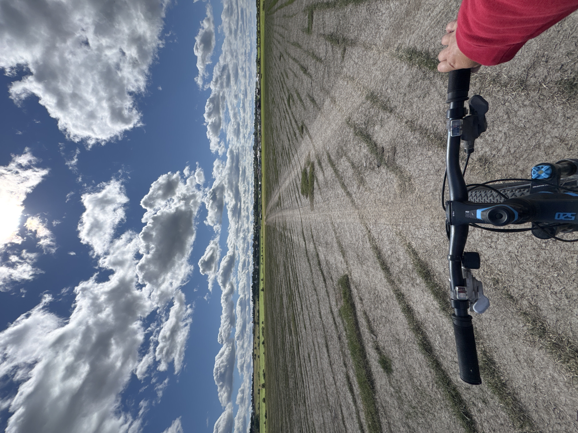 The image shows a first-person perspective of someone riding a bicycle on a dirt path through a field. The field appears to be recently tilled or planted, with visible rows of dry earth and sparse green growth. In the distance, there is a line of trees and buildings, suggesting a settlement. The sky is bright blue with scattered white clouds, and the sun is visible, creating lens flare. The bicycle's handlebars and a person's hand in a red jacket are in the foreground. The scene is likely located in Kempele, Russia, given the context provided, implying a rural or semi-rural agricultural landscape.