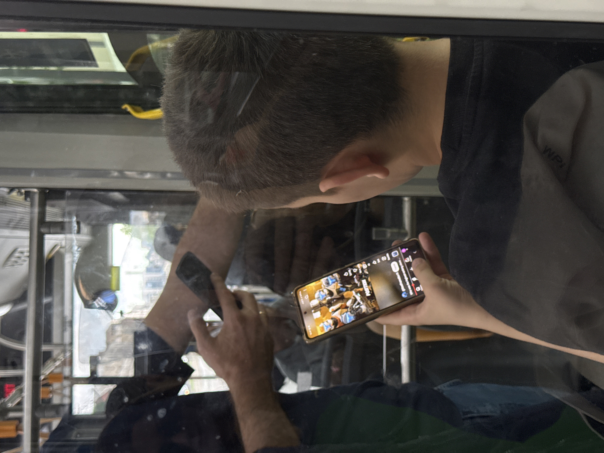 The image shows a person on what appears to be a public transport vehicle, possibly a bus, as indicated by the window frame and the glimpse of the vehicle's interior and exterior. The person is holding a smartphone, and the screen displays a video or social media feed. The reflection in the window shows the surroundings, which may be a city street with buildings and vegetation. While details are limited, the reflection does not allow for precise location determination within Belgrade, Serbia.