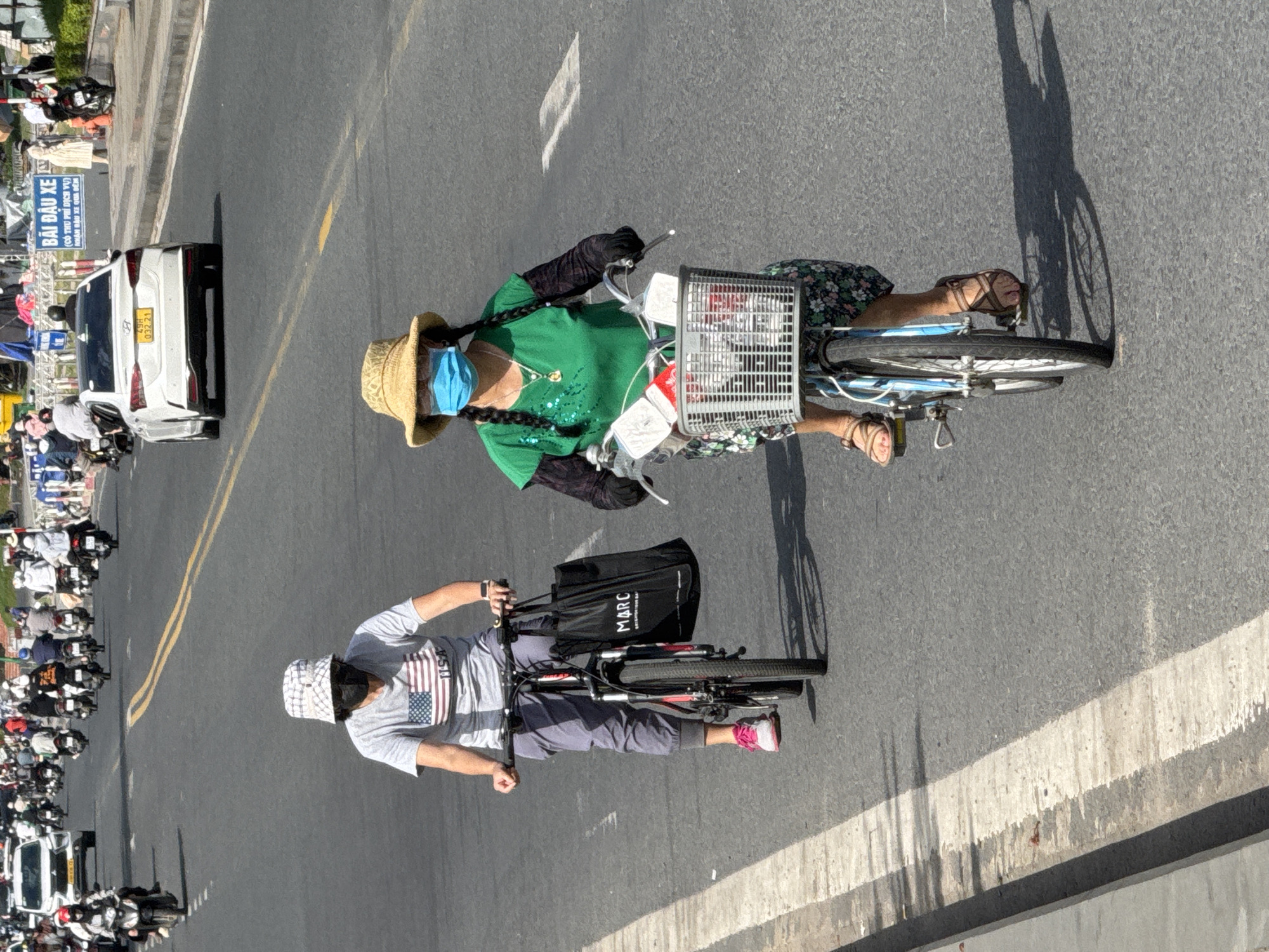 In Da Lat, Vietnam, two individuals are riding bicycles on a paved road. The foreground features a person wearing a straw hat, a green shirt, and a blue face mask, riding a bicycle with a basket containing items. To their left, another person is riding a bicycle while carrying a black tote bag. Both individuals are in motion, navigating a traffic-filled street with cars and motorcycles in the background. The lighting suggests it is daytime, with shadows cast by the riders and their bicycles. The road is marked with white and yellow lines.