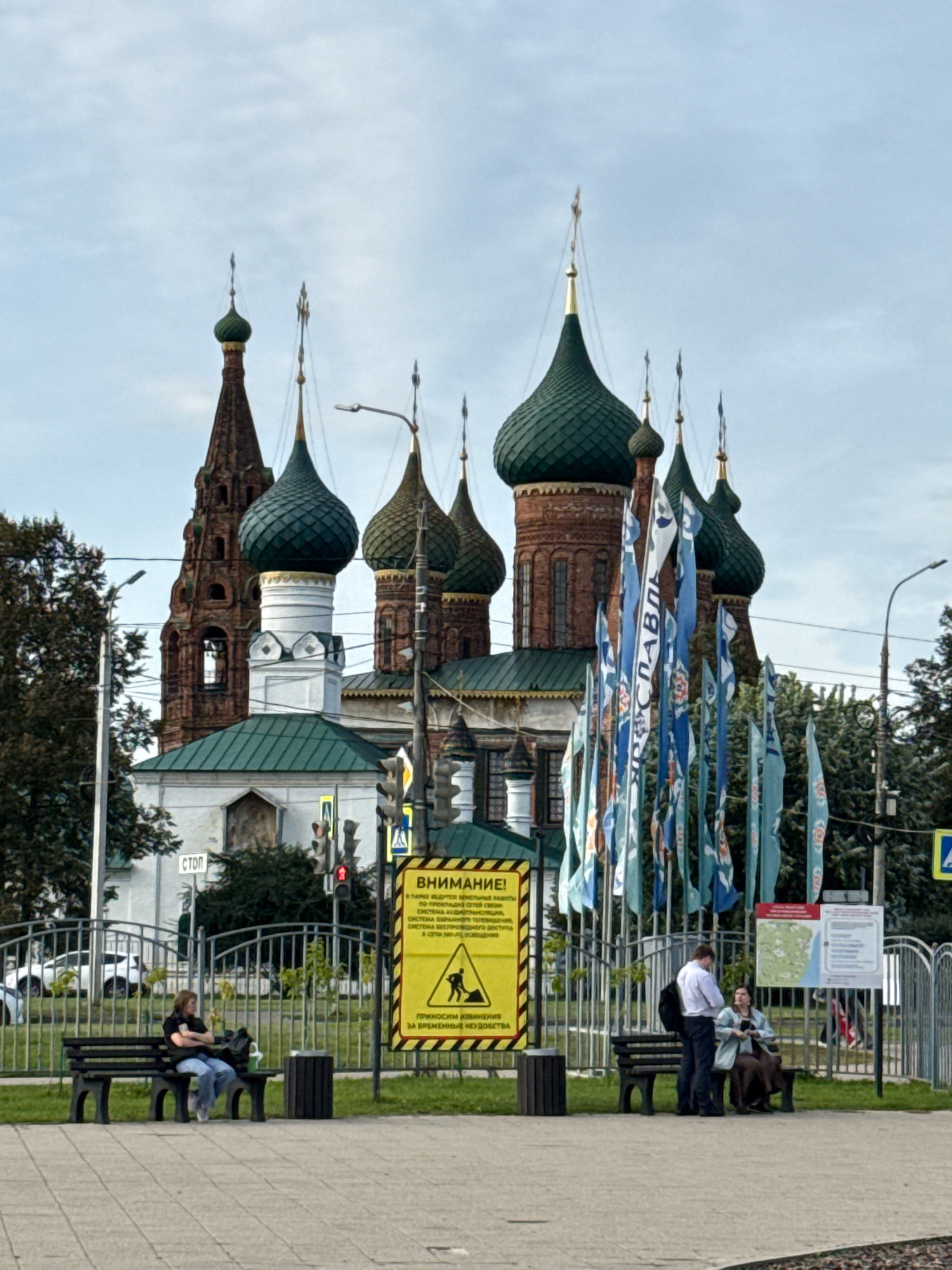In Korobovo, Russia, a wide-angle, eye-level shot captures a religious building with multiple onion domes and a tall, slender bell tower. Flags with blue and white designs are displayed in front of the building. In the foreground, two individuals are seated on a park bench, while another pair stands nearby. To the right of the scene, a yellow warning sign with a graphic illustration of a person working is visible. A metal fence encloses a grassy area behind the bench. The sky is overcast.