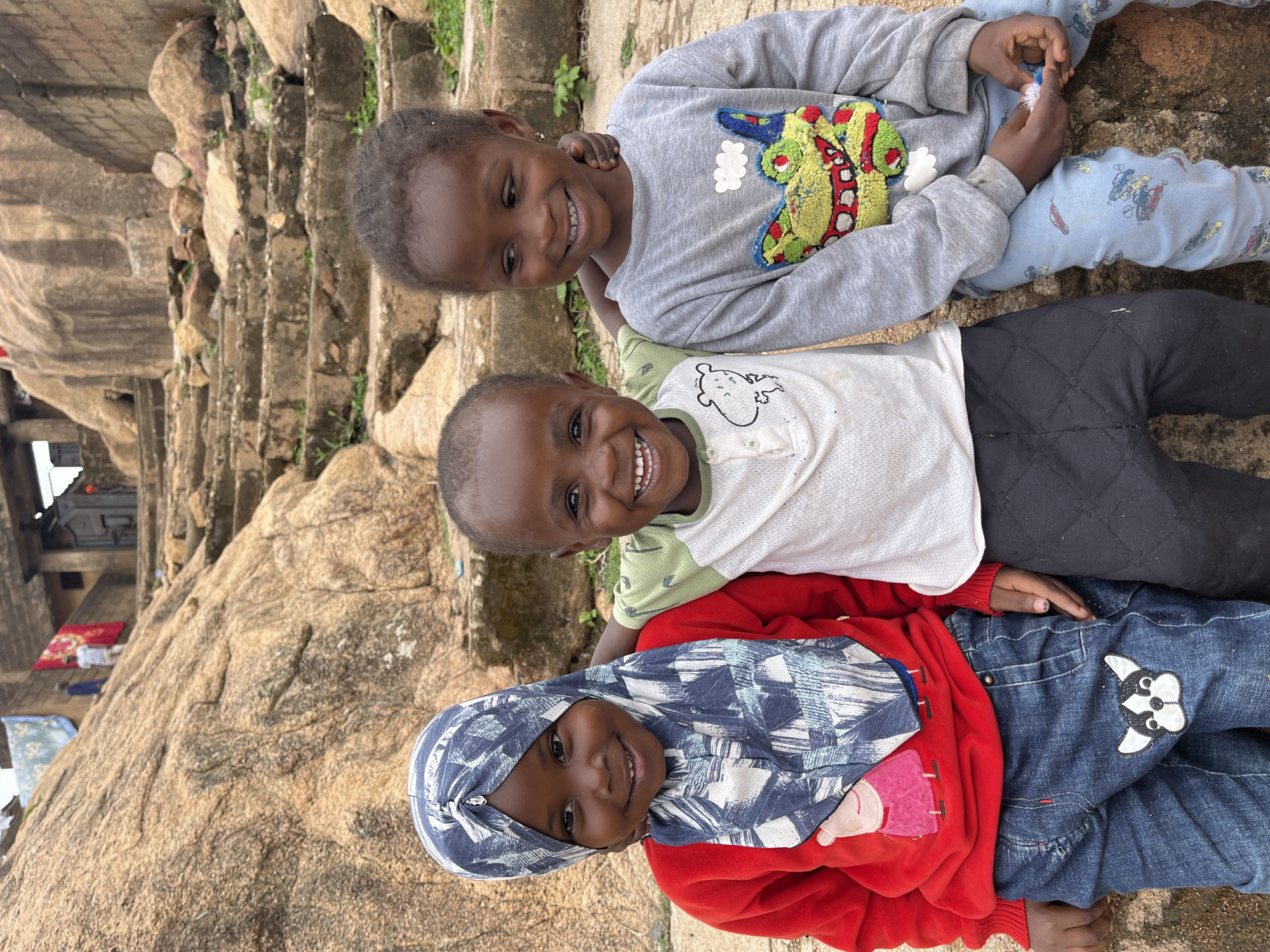 Three smiling children are lying on a rocky outcrop, stacked vertically, appearing to be posing for the camera. The top child is wearing a grey sweater with a cartoon monster and blue patterned pants. The middle child wears a white t-shirt with a simple animal graphic and black pants. The bottom child wears a red hooded sweatshirt with a patterned headscarf, and blue jeans with a dog graphic on the leg. The children are positioned in front of a large rock formation with a building and steps visible in the background. The scene takes place in the city of Gura Top, Nigeria.