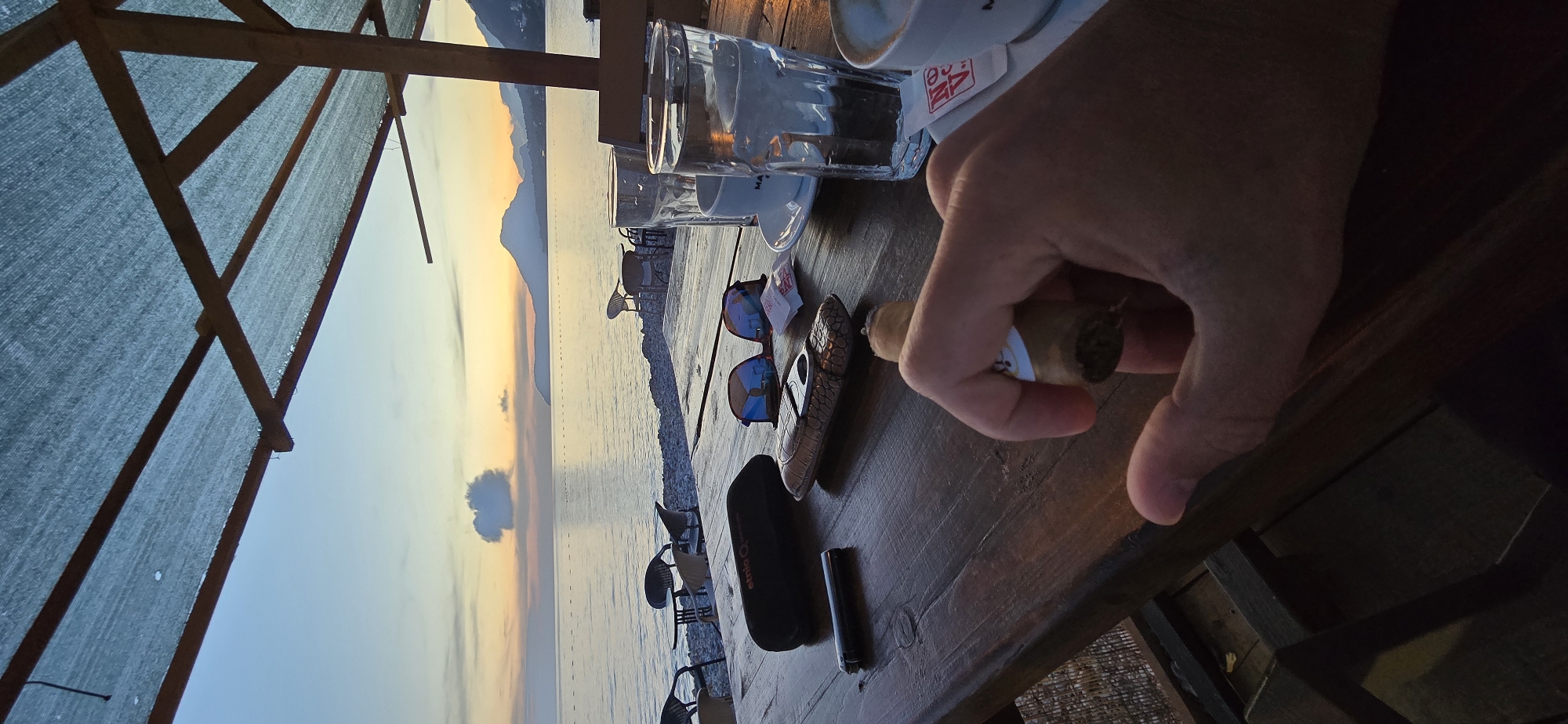 This vertically oriented image captures a serene sunset scene at a beachside establishment in Bar, Montenegro. In the foreground, a person's hand holds a cigar, which is about to be lit, indicated by the nearby black cylindrical lighter and an alligator-skin textured cigar cutter resting on a wooden table. A black cigar case, partially revealing the text "Optic Vue," and a pair of mirrored sunglasses with colorful reflections are also on the table.
Two glasses of water and two white ceramic coffee cups on saucers suggest beverages are being enjoyed. Small white paper packets, possibly sugar or napkins, are visible, one with a partial "MAT..." and a red, possibly "AA" or similar, logo. Overhead, a light blue fabric canopy, supported by wooden beams, provides shade.
Beyond the table, the calm Adriatic Sea stretches towards the horizon, reflecting the warm, golden, and orange light of the setting sun. Distant mountains rise from the far shore, silhouetted against the vibrant sky, which also features a few soft clouds. Several empty beach chairs or sun loungers are visible on the pebbly shore, hinting at a quiet and relaxed atmosphere as dusk settles. The scene evokes a sense of peaceful leisure and contemplation.