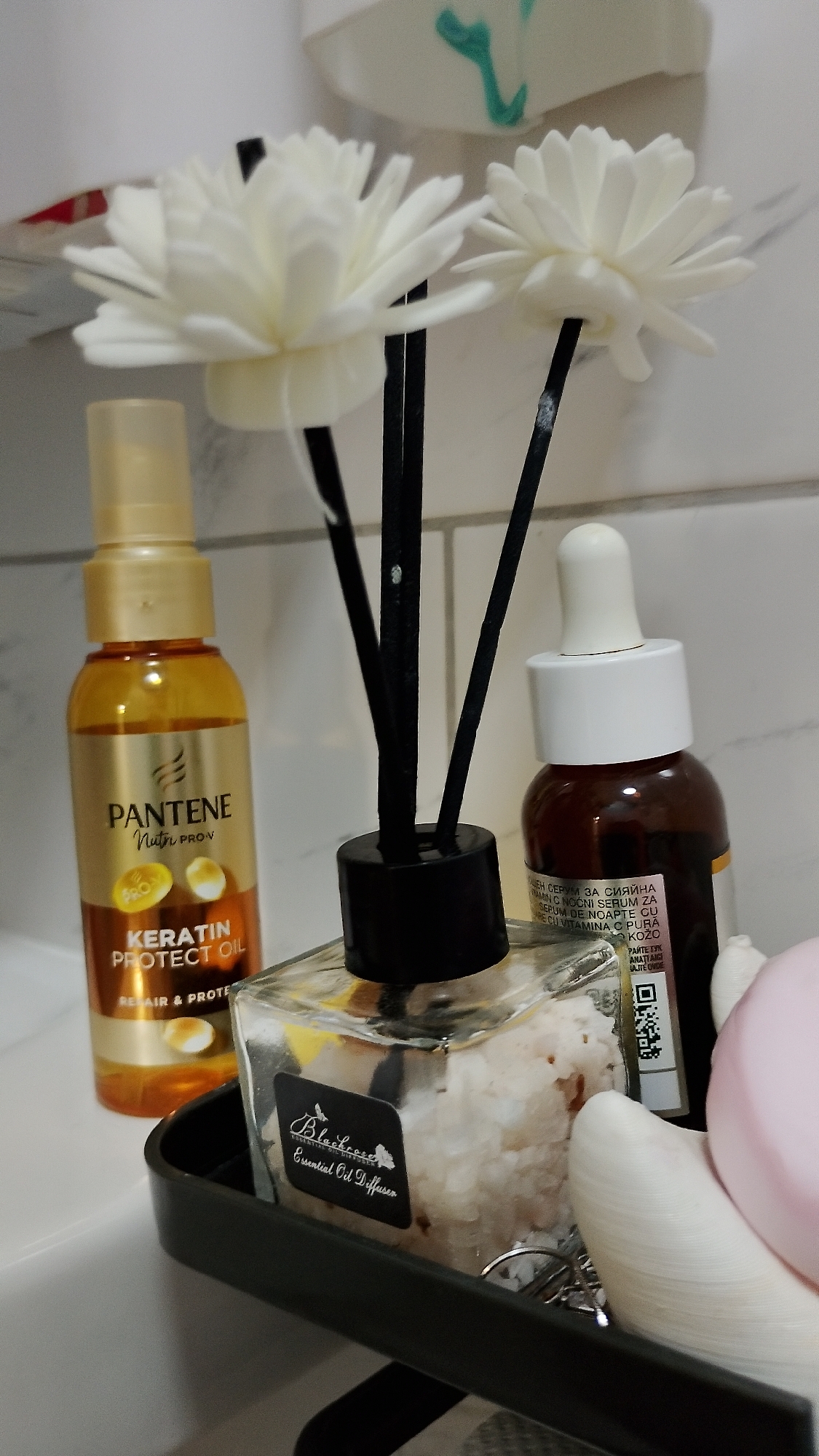 The image depicts a close-up, eye-level view of a bathroom counter, specifically on a tiled surface, in Smolyan, Bulgaria. The foreground is dominated by a black plastic tray containing several personal care items. Prominently featured is a glass diffuser with a black lid and three black reeds, filled with clear liquid and white crystal-like fragments. To the left of the diffuser is a gold-colored plastic bottle of Pantene Keratin Protect Oil. To the right of the diffuser is a small brown glass bottle with a white dropper cap, containing a dark liquid. The label on this bottle has some visible text, including Cyrillic script and a QR code. In the background, two white decorative flowers on thin black stems emerge from the diffuser. Further back, a white wall with a faint teal-colored graphic is visible. The lighting appears to be artificial, common for an indoor bathroom setting. No people are present in the image, and the focus is entirely on the arrangement of these products.