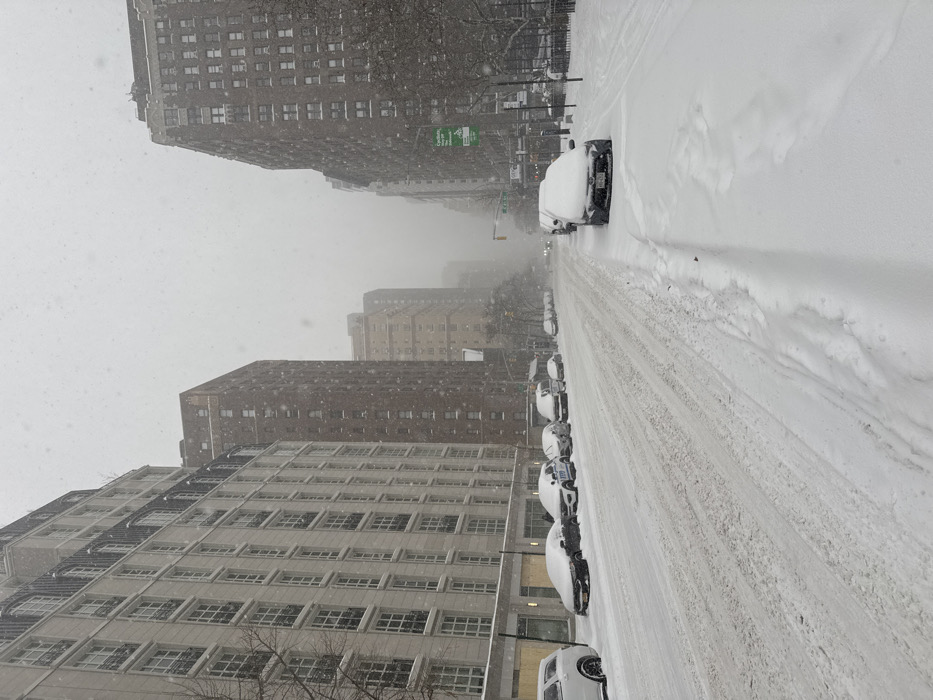 An urban street during a heavy snowfall. Buildings line both sides of the street, and cars are parked along the curb, mostly covered in snow. The street is coated with a thick layer of snow, and visibility is reduced due to the ongoing snowfall. Snowflakes are visible in the air. The scene appears to be in a city, possibly New York City, given the architectural style.
