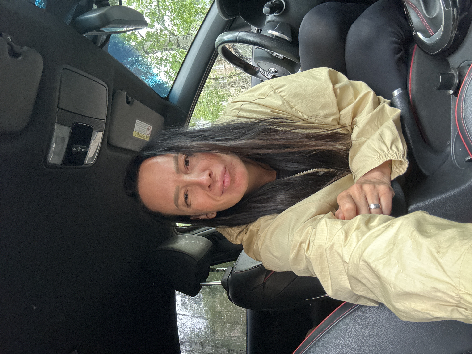 A woman is taking a selfie in the driver's seat of a car. She is wearing a light yellow jacket. The car interior is black. Outside, through the windshield, trees are visible and it appears to be raining. The location is Chaikovsky, Russia.