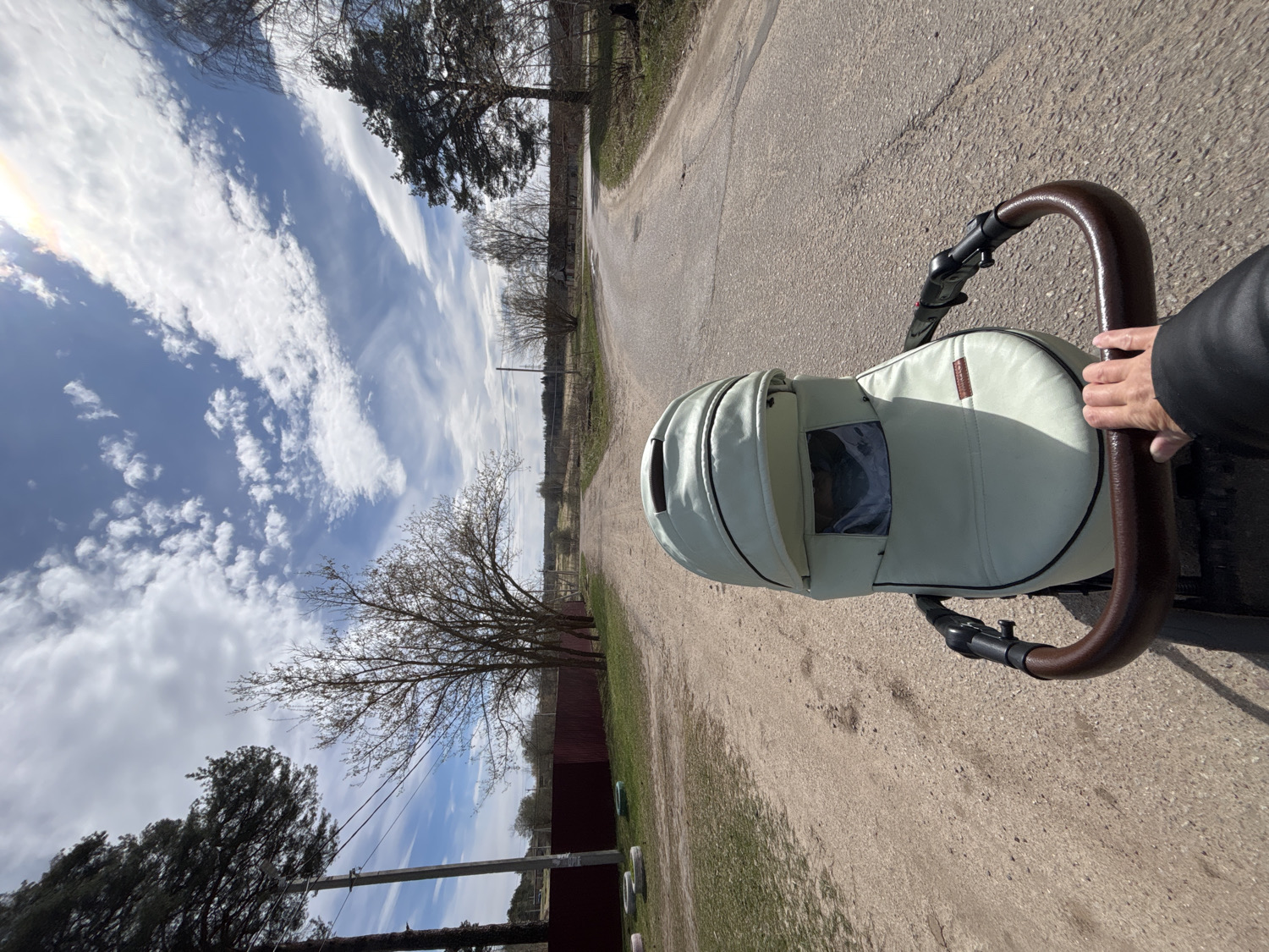 An outdoor, eye-level shot, taken from a first-person perspective, captures someone pushing a baby stroller along a dirt or gravel road in Раздолье, Russia. The image is rotated counter-clockwise, showing the sky on the left and the ground/road on the right.
In the foreground, a hand, wearing a black jacket sleeve, grips the dark brown, curved handle of a baby stroller. The stroller itself is light-colored, possibly mint green or cream, with dark trim and a small, rectangular label on its side, the text of which is illegible. The stroller's canopy is partially extended, and a clear plastic window is visible on its side.
The road ahead is unpaved, appearing as a light brown, earthy path stretching into the distance. Bare trees with thin branches line both sides of the road, suggesting an early spring or late autumn season. Some darker evergreen trees are also visible. In the background, there's a hazy horizon line with more distant foliage and what appear to be power poles or fences.
The sky is a prominent feature, filled with varied clouds against a bright blue background. There are long, wispy, white clouds stretching across the sky, as well as some smaller, puffier clouds. The sun is visible in the upper left, casting a bright lens flare and indicating clear, daytime weather. The overall impression is one of a peaceful, possibly rural or semi-rural stroll under a bright sky.