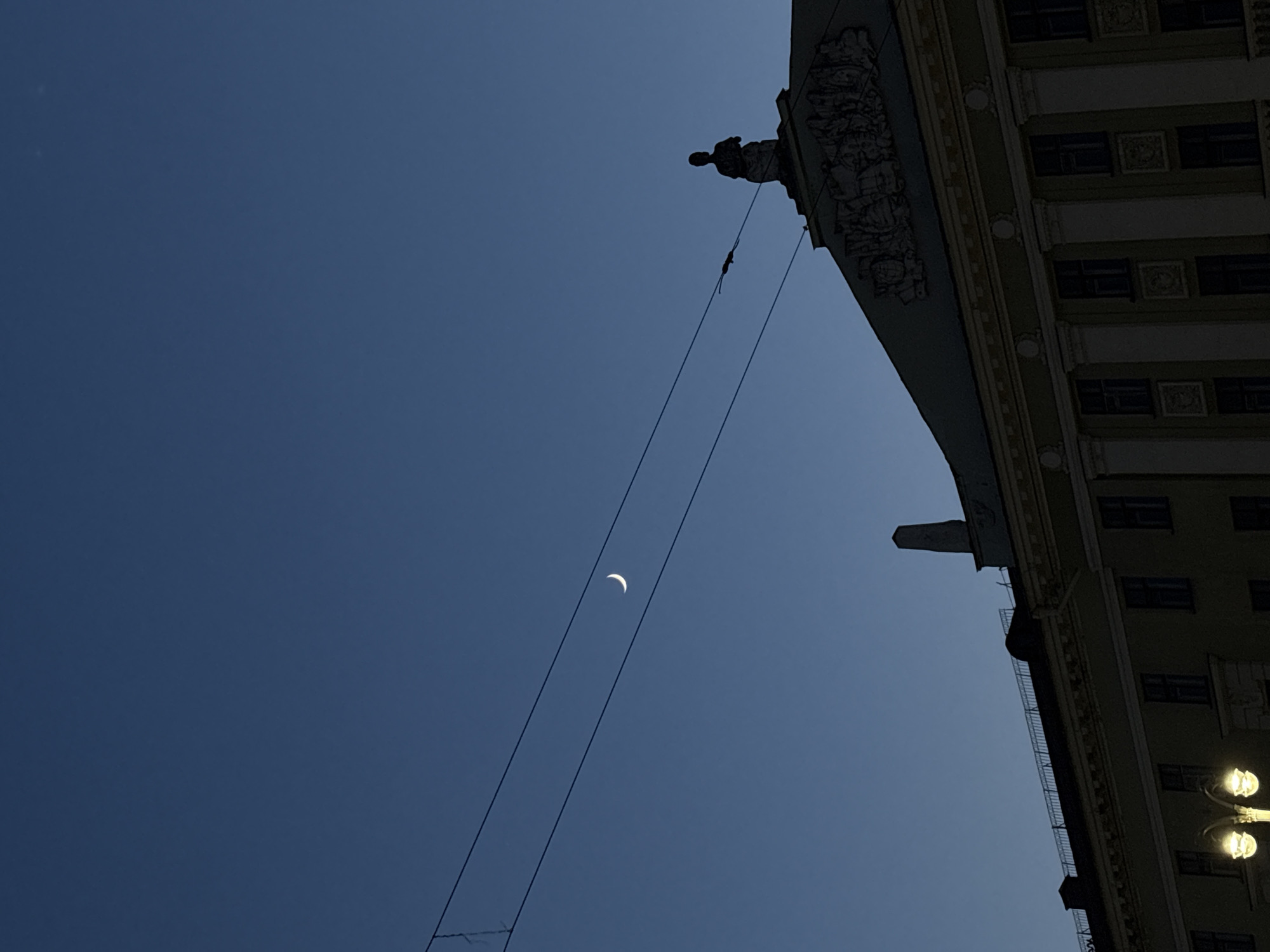 The image shows a low-angle view in what is likely Minsk, Belarus. In the lower left of the sky a thin crescent moon is visible. Two overhead wires extend vertically upwards through the moon. The right side of the frame is dominated by the corner of a large building. The building is ornate, with classical architectural details visible on its façade, including sculptures along the roofline and decorative features around the windows. A streetlight is visible at the bottom right corner of the building. The overall lighting indicates dusk or early evening. The sky is a deep blue. The building is mostly in shadow. No people are visible.