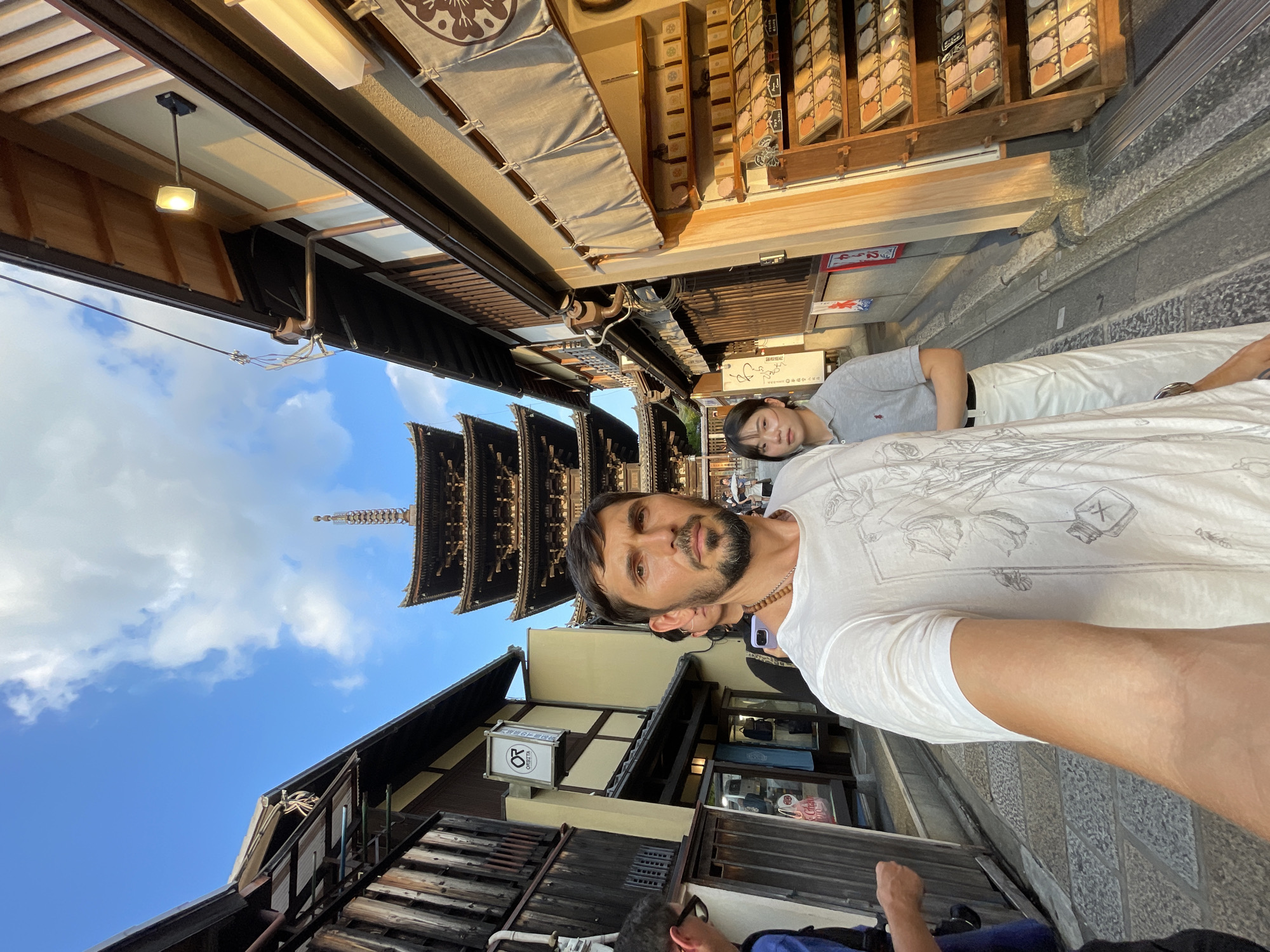 The image depicts a street scene in Kyoto, Japan, with a prominent five-story pagoda in the background. In the foreground, a man is taking a selfie, holding the camera out towards himself. He is wearing a white t-shirt with a floral design. To his right, a woman in a grey polo shirt and white pants stands looking forward. The street is lined with traditional Japanese wooden buildings, one of which has a shop displaying various items on shelves. The sky is blue with scattered clouds, and the lighting suggests it is late afternoon. The architecture and the pagoda strongly indicate the location is within the Higashiyama District, possibly near Kiyomizu-dera Temple or Yasaka Pagoda.