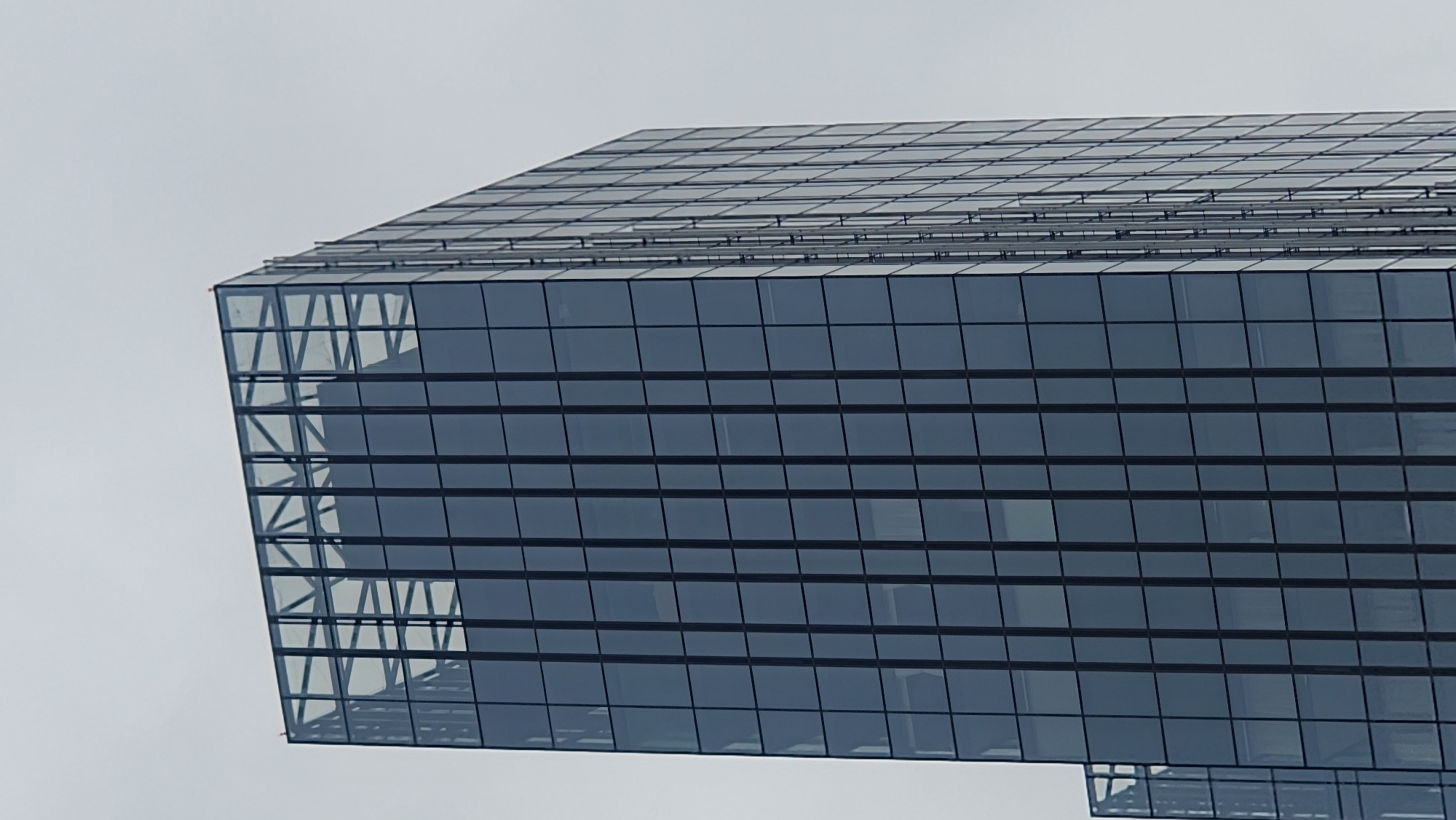 The image shows a close-up, low-angle view of a modern skyscraper with a glass facade. The building's geometric design is emphasized, with clean lines and a grid-like pattern of windows. The top surface of the building appears to be flat and covered in panels, possibly solar panels or a rooftop structure. The side of the building features a sharp, angular corner that extends outwards, revealing a structural framework behind the glass. The glass itself is tinted dark blue or grey, reflecting the overcast sky. The scene is dominated by the building's architecture, with a pale, featureless sky filling the background, suggesting a cloudy or hazy day. There are no people or discernible activity visible in the frame. The overall impression is one of architectural modernity and scale, with the building appearing detached from its surroundings due to the lack of context. The location context is Moscow, Russia, which implies this is likely a contemporary office building or residential complex in the city. No text is visible in the image.