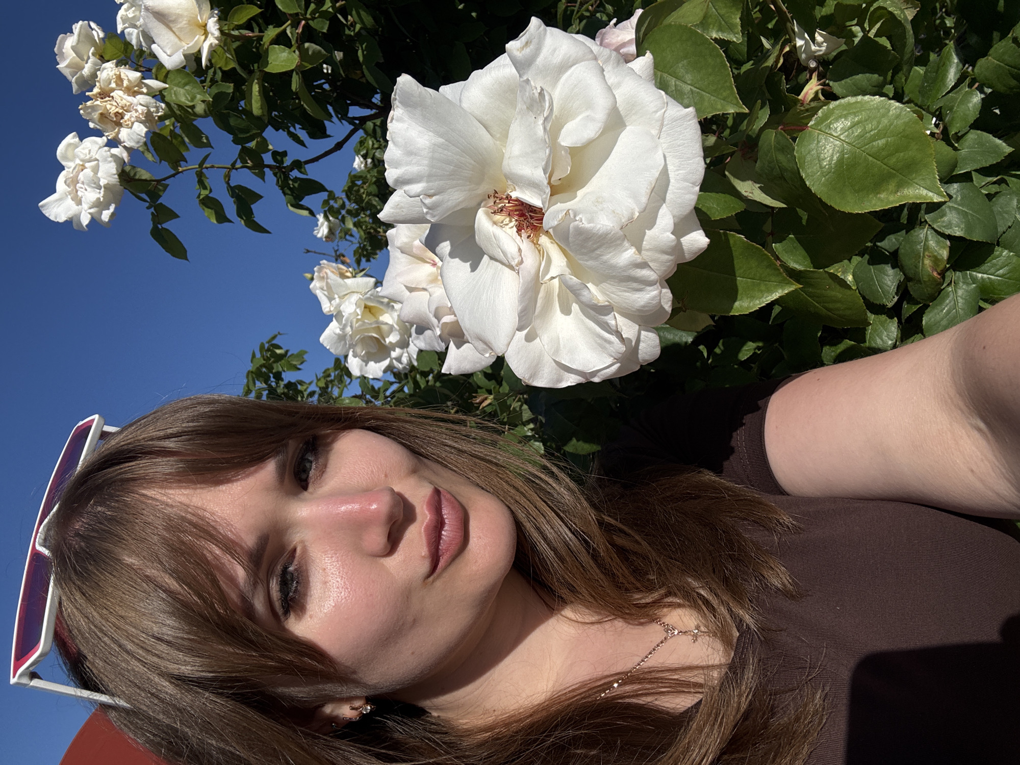 A young woman with brown hair and brown clothing is positioned in front of a white rose bush under a clear blue sky. She appears to be taking a selfie, as her arm is visible on the right side of the frame. A pair of sunglasses is perched on her head. The scene does not provide enough detail to determine whether this image was taken in город Ялта, Ukraine.