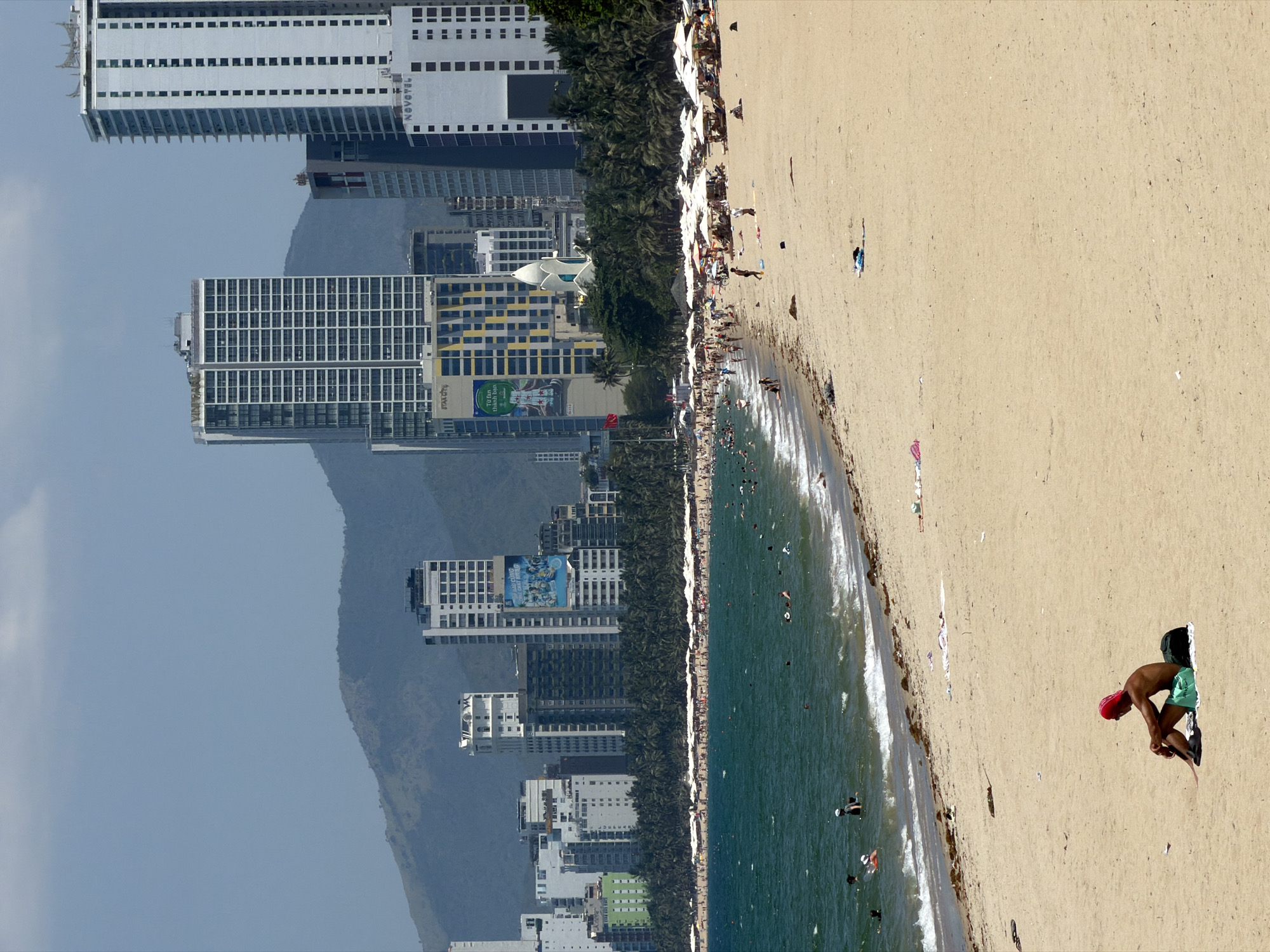 This image captures a panoramic view of a bustling beach in Nha Trang Ward, Vietnam. The scene is set under a light blue sky with scattered faint clouds, overlooking distant, hazy green mountains that form the backdrop.
The city skyline, situated behind a dense line of palm trees, features numerous modern high-rise buildings. Notable structures include a building with "VINI BAN" partially visible, another marked "STAR CITY" with yellow and black patterned sections, and a prominent white "NOVOTEL" tower. A distinctive white, lotus-shaped architectural feature is integrated into the urban landscape among these buildings. A large billboard depicting people swimming is visible on one of the mid-rise structures.
The beach itself is a wide stretch of light golden-brown sand. In the mid-ground, a significant number of people are gathered on the beach, with many relaxing under a uniform arrangement of white umbrellas. Other individuals are seen walking along the shoreline.
The deep blue-green ocean occupies the left side of the mid-ground, with gentle waves breaking near the shore. Numerous individuals are swimming, wading, and floating in the water. Some dark organic debris is visible along the waterline.
In the immediate foreground, a lone individual wearing a red head covering and dark shorts is seated directly on the sand, facing the ocean. A dark backpack and a light green towel or mat are positioned beside them.