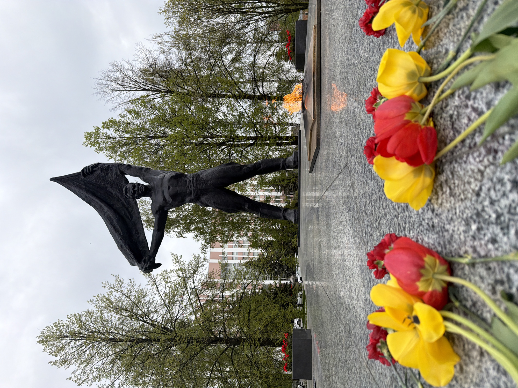 The image shows a monument and eternal flame in Izhevsk, Russia. The statue is of a muscular man holding a banner. The eternal flame burns behind the statue on a gray stone platform. Red and yellow tulips are laid on the platform in the foreground. Behind the monument are trees with new spring growth. In the background, a multi-story building can be seen. The sky is overcast.