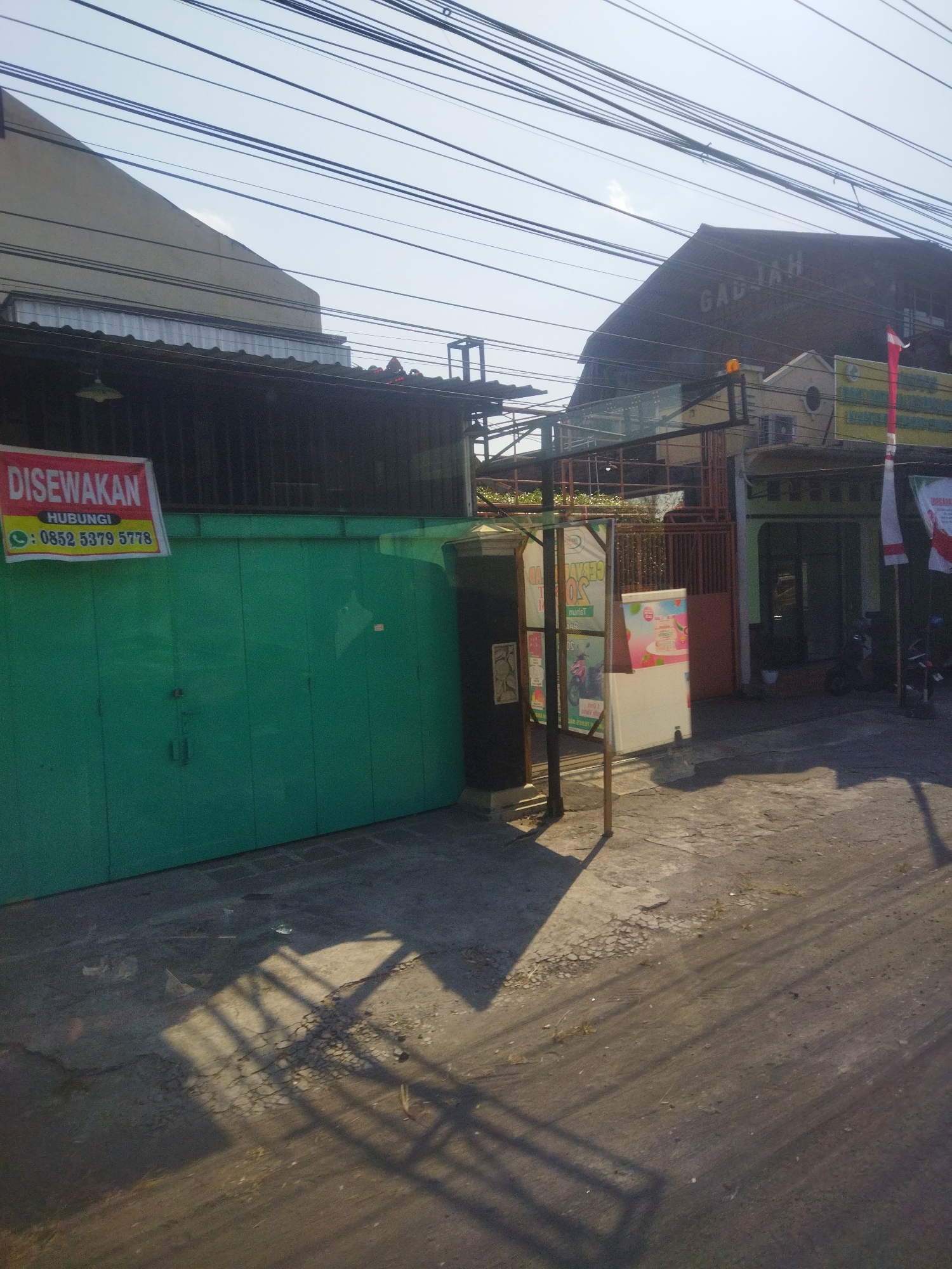 The image shows a street scene in Begajah, Indonesia. On the left is a building with a green metal roll-up door, and a sign above it reads "DISEWAKAN" (For Rent) with a phone number. To the right of the green door is a black cylindrical object, likely a trash bin or storage container. Further to the right are several buildings with various signs and awnings. One building has the word "GADJAH" written on its roof. A red and white flag is visible, along with an Indonesian flag. The ground is paved and appears to be a road or sidewalk. Power lines crisscross the sky. The scene is lit by sunlight, casting shadows across the ground. No people are visible in the image.