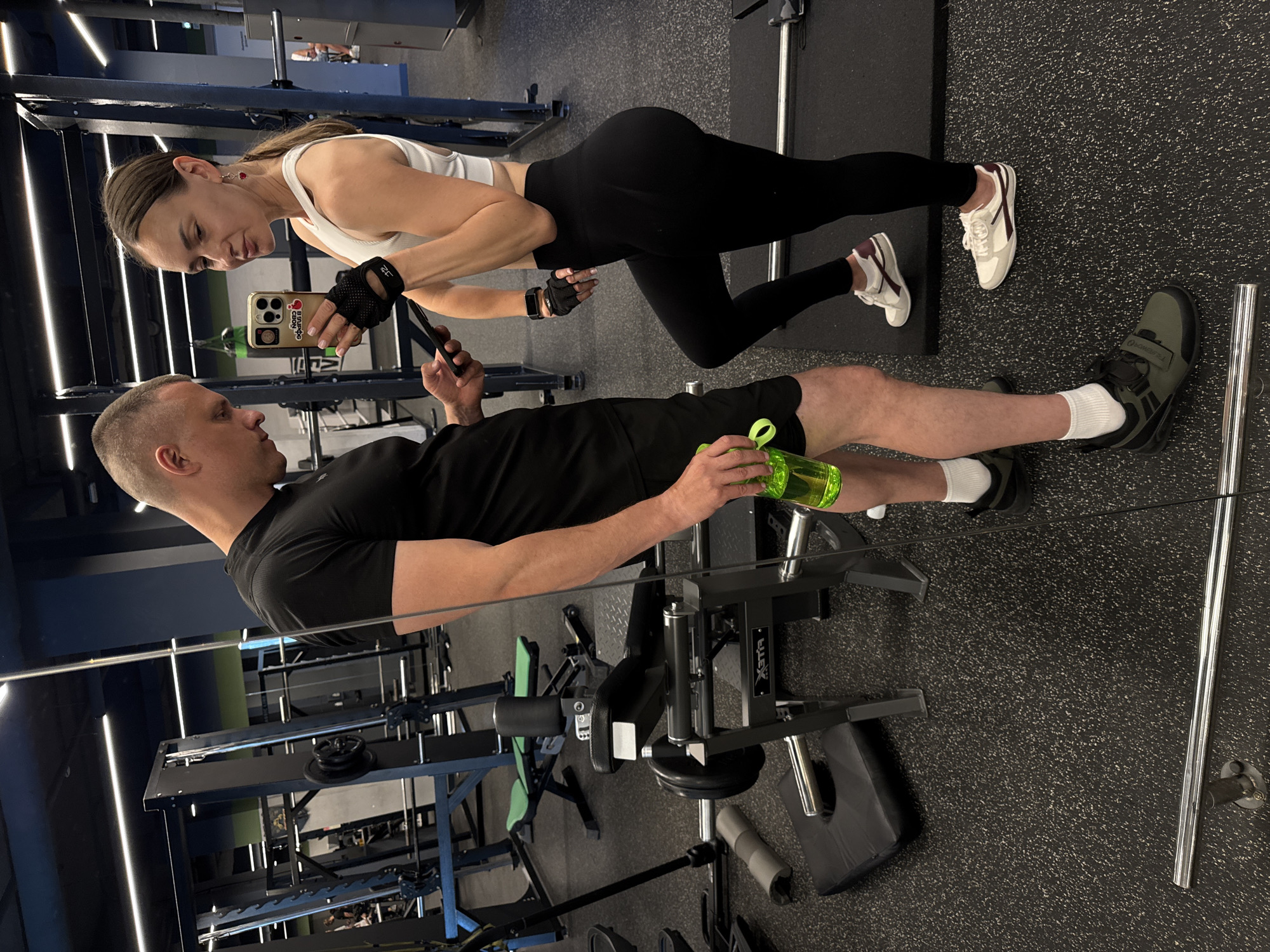 The image shows a man and a woman in a gym in Sochi, Russia. The woman is wearing a white sports bra and black leggings, and the man is wearing a black t-shirt and shorts. The woman is holding a smartphone and appears to be taking a selfie, while the man is looking at his phone. They are both dressed in athletic wear, suggesting they are working out. The gym appears to be well-equipped with various exercise machines. The lighting is bright, indicating it could be daytime or the gym has good artificial lighting. The overall mood seems casual and focused on fitness. There is no visible text in the image.
