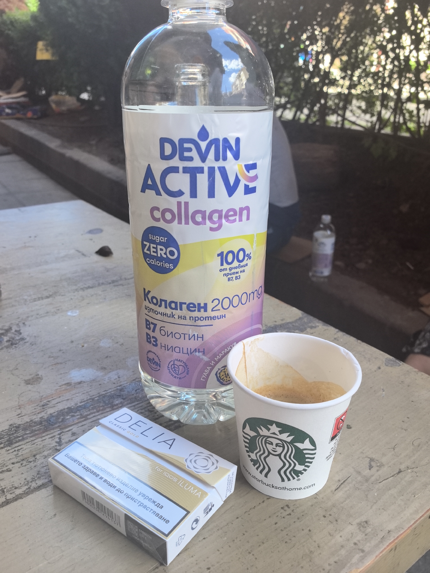 Here is a forensic description of the image:
**Scene:** A slightly weathered, light-colored wooden surface.
**Objects:**
* A clear plastic bottle of "Devin Active Collagen" water, with prominent labels in Bulgarian and English. The bottle is mostly full.
* A white Starbucks paper cup containing a dark brown liquid (presumably coffee). A filter is inside the cup.
* A white pack of "Delia Classic Gold" cigarettes, labeled as "for IQOS ILUMA". There are warnings in Bulgarian on the pack.
**Environment:**
* The scene appears to be outdoors, with greenery visible in the blurred background.
* There are people in the background, but they are out of focus.
**Additional Details:**
* The location is inferred from the use of Bulgarian language on the products.
* The brand names and packaging details are clearly visible.
