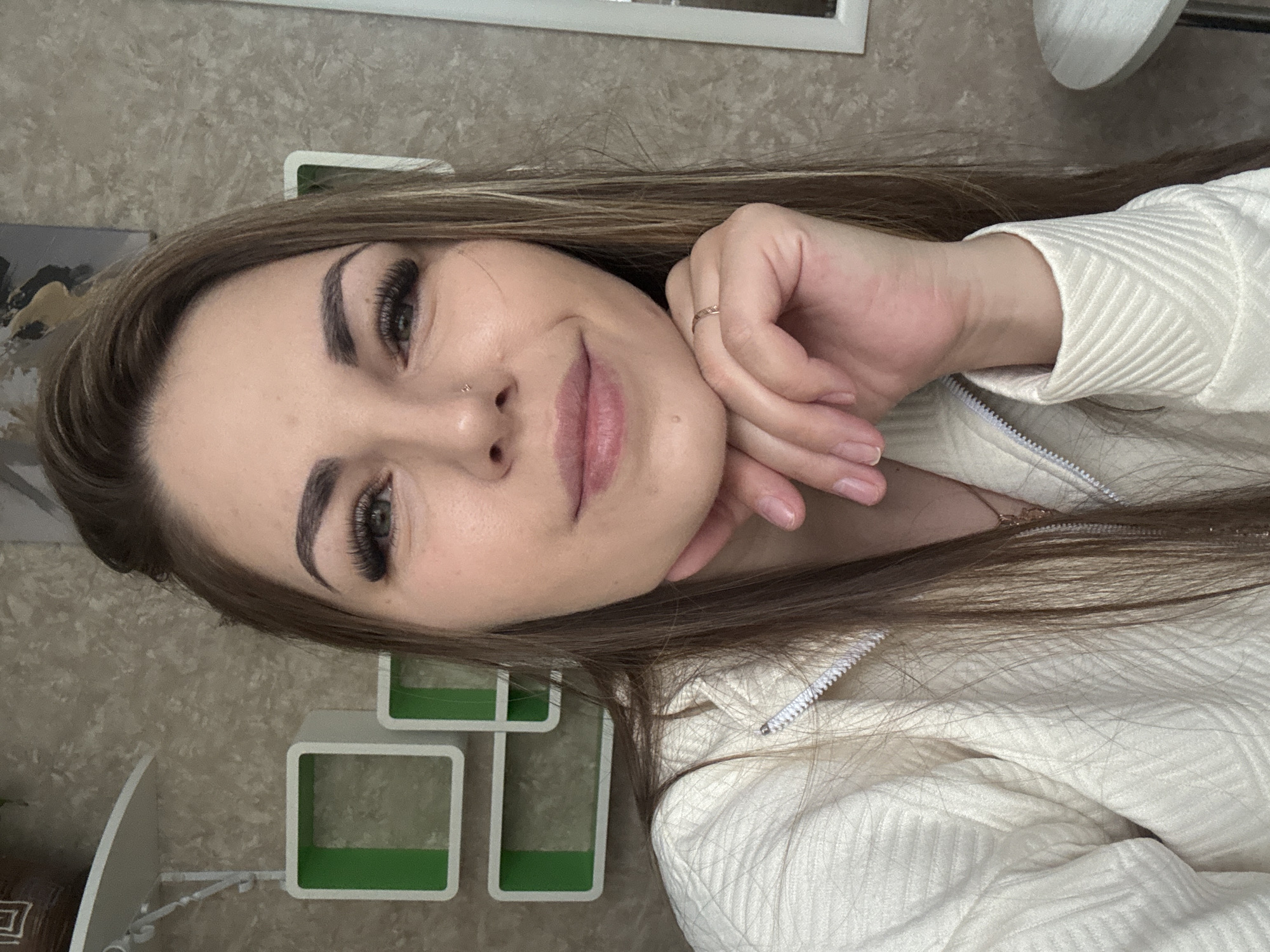 A person is depicted in a selfie-style portrait. The individual has long brown hair and is wearing a white jacket with a zipper. The person's hand is resting on their face. The background includes a patterned wall, some square shelving units with green accents, and a light fixture. It is not possible to ascertain from the image the location of Razdolie, Russia.