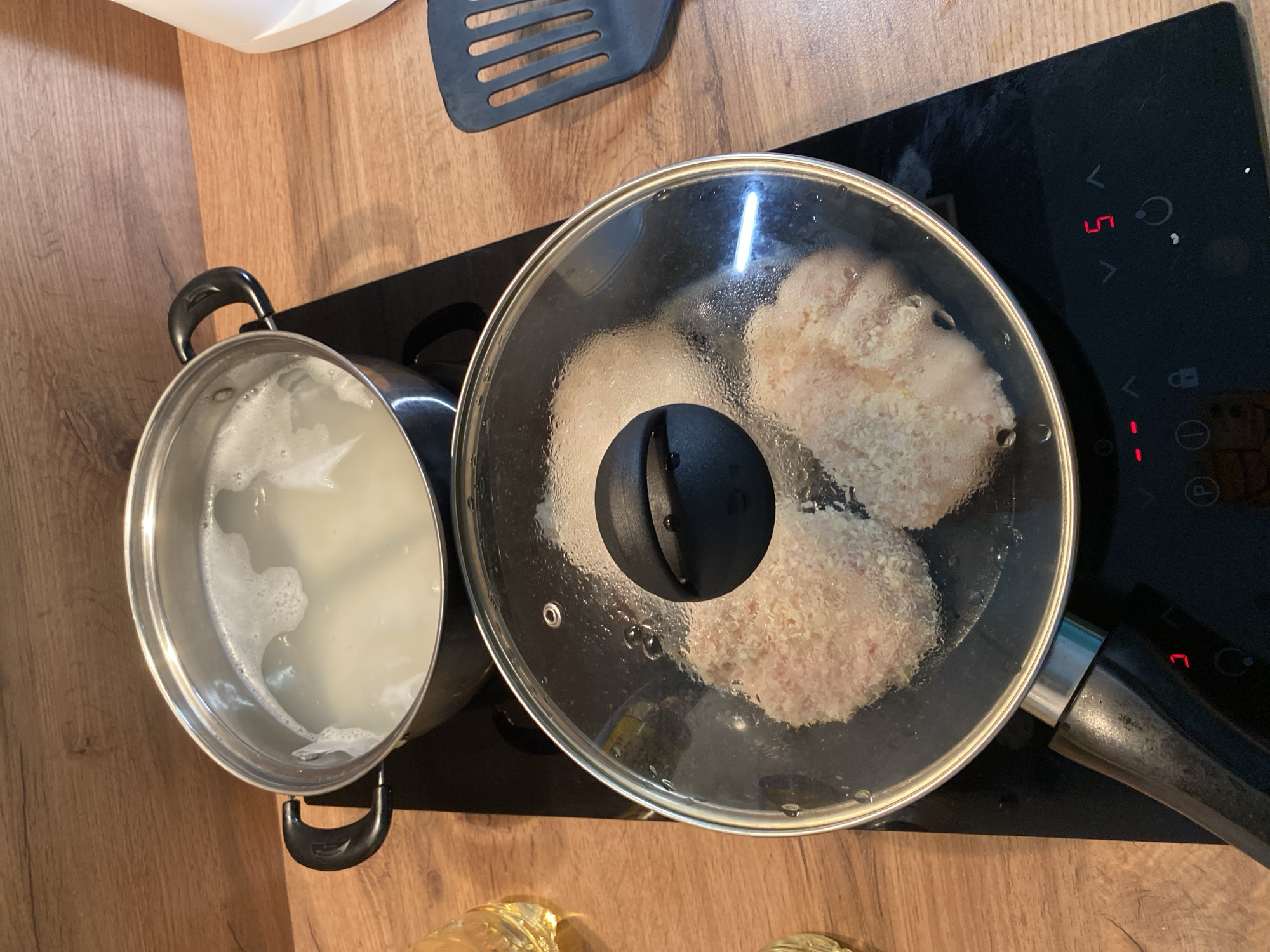 In Yekaterinburg, Russia, on a kitchen countertop with a wooden finish, a close-up shot captures two cooking activities. In the foreground, a frying pan with a glass lid is placed on a black induction stovetop. Inside the pan, two breaded food items, likely cutlets or patties, are being cooked. Steam has condensed on the underside of the glass lid, indicating the food is cooking. The stovetop displays a digital temperature setting of '5' in red. Behind the frying pan, a stainless steel pot with a black handle is also on the stovetop, containing boiling water or a light-colored liquid with some frothy bubbles. To the right of the stovetop, a black slotted spatula rests on the countertop. A glass with a yellow liquid is partially visible on the left edge of the frame. The lighting suggests an indoor, domestic setting. No individuals are present in the scene.