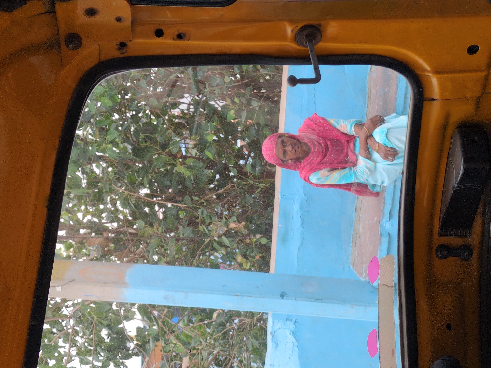 An elderly woman, wearing a pink headscarf and a light blue and pink outfit, sits on a ledge. Her arms are crossed in front of her. She is viewed through the passenger side window of a yellow auto-rickshaw, with the yellow interior of the auto-rickshaw framing the lower part of the image. The woman is seated in front of a bright blue wall, with green foliage visible in the background. The scene appears to be in Jodhpur, India, based on the auto-rickshaw and the style of dress.