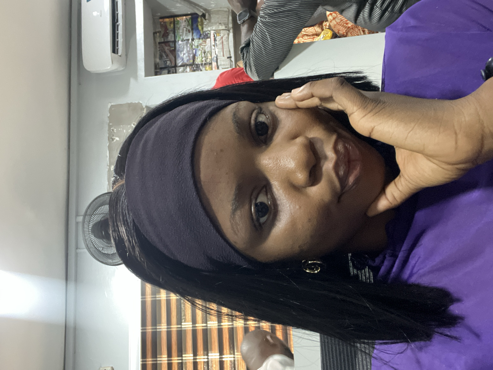 A woman with dark hair and a dark purple headband is looking at the camera with her right hand touching her cheek. She is wearing a dark purple shirt. In the background are a fan, an air conditioner, a window with blinds, and a magazine rack. The location is likely in Ikeja, Nigeria. Other people are partially visible in the background.
