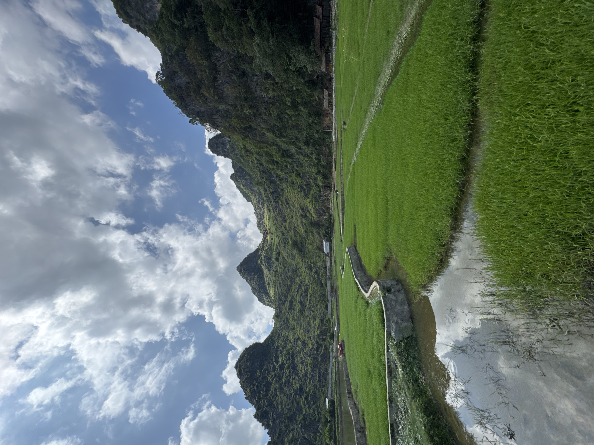 The image portrays a landscape in Đồng Văn Commune, Vietnam, featuring rice paddies adjacent to a limestone mountain range under a partly cloudy sky. The rice fields display vibrant green foliage and are separated by narrow irrigation canals. The mountains are covered in dense vegetation, with occasional structures visible along their lower slopes. The sky features cumulus clouds and patches of blue. A water surface reflecting the sky and landscape is present on the right side of the frame.