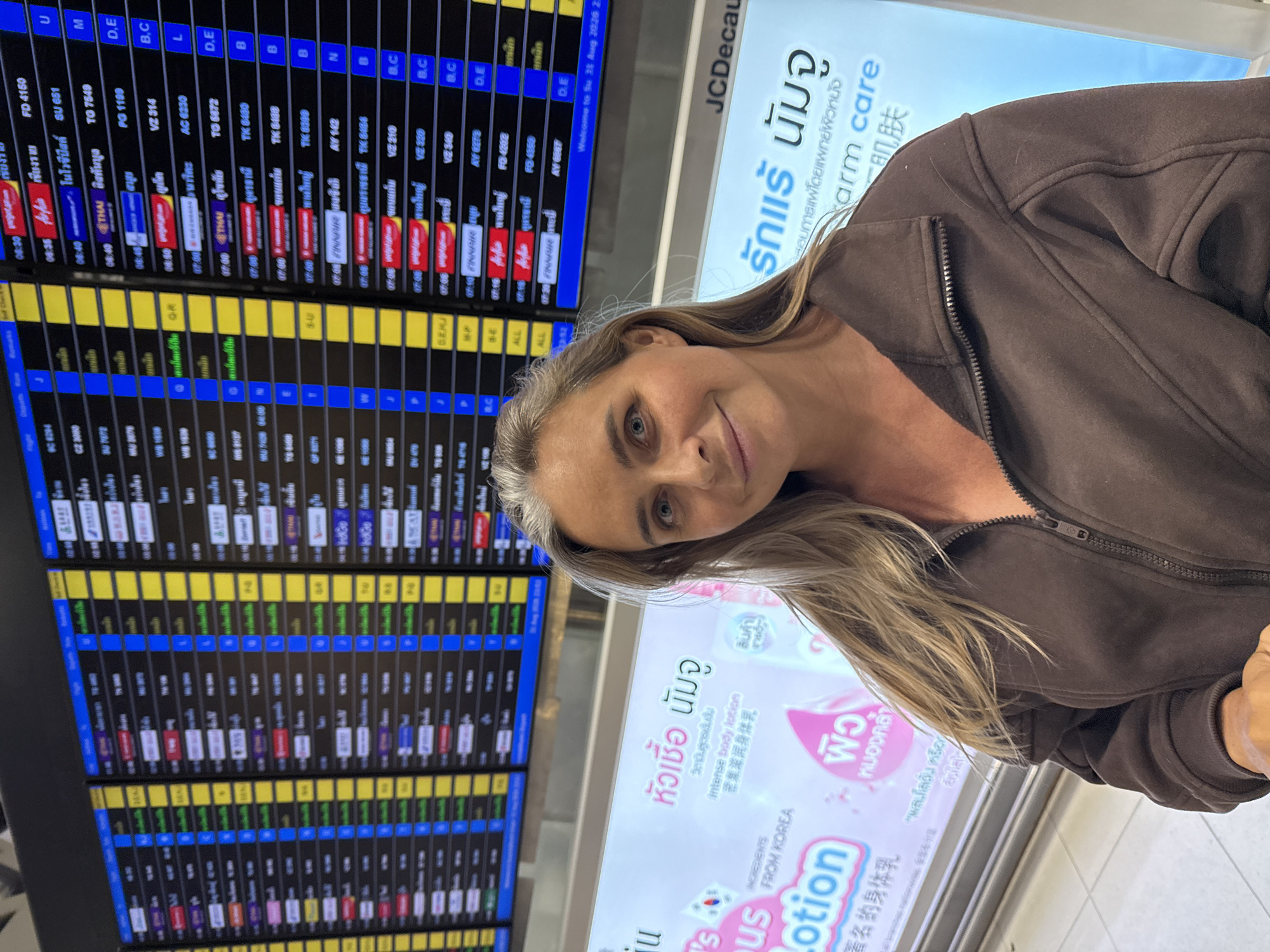 A woman is posing for a selfie in front of a departure board at an airport in the city of Nong Prue Subdistrict, Thailand. The departure board shows flight information, including flight numbers, destinations, and times. The woman is wearing a brown zip-up hoodie. In the background, there is a digital advertisement displaying information in Thai and English, including "Intense body lotion" and "arm care". The overall scene suggests a travel environment.