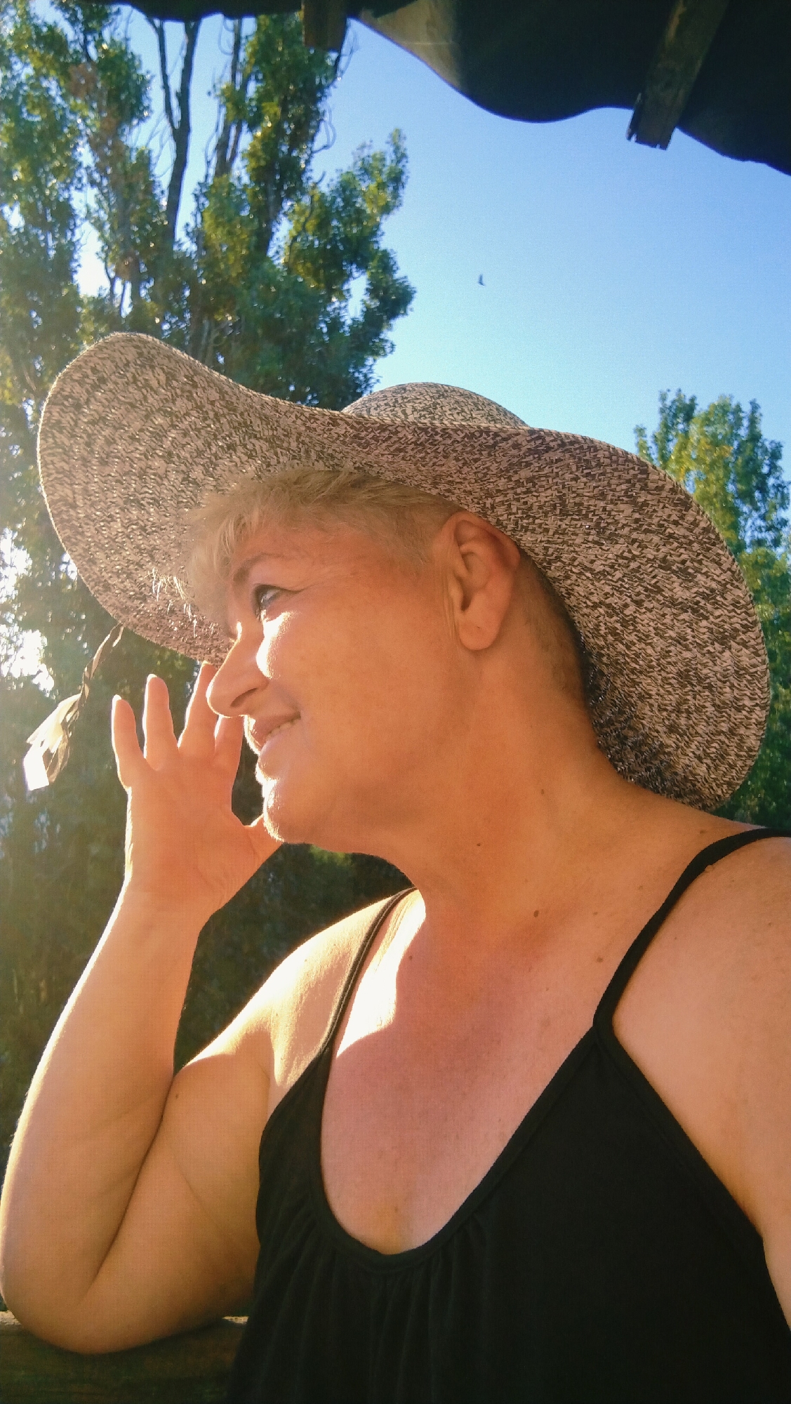 An outdoor, close-up, head and shoulder shot captures a woman wearing a wide-brimmed, textured hat and a black camisole. She is positioned in profile, facing left, with her right hand raised towards her face. The background consists of green trees and a clear blue sky, with a dark structure visible overhead. The lighting suggests a sunny day, with sunlight illuminating the woman's face and arm, casting shadows on her chest and neck. The image appears to be taken from a low angle. There is no discernible information about the location in the city of Horishni Plavni, Ukraine within the provided image.
