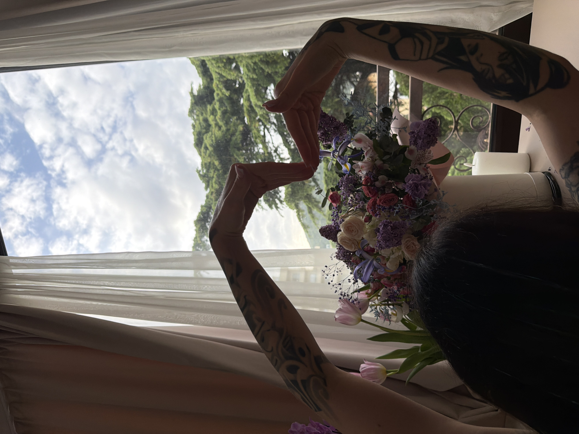 The image presents a close-up, intimate view from an indoor setting looking out a window in Budva, Montenegro. The primary subject is a person, seen from a high angle, with their tattooed arms raised and hands forming a distinct, intertwined shape, possibly framing the view or the flowers. Visible tattoos adorn both arms, featuring intricate designs including what appears to be a stylized face on the right arm. The back of the person's head, with dark hair, is seen in the foreground to the right.
Immediately in front of the person is a large, colorful bouquet of various flowers. The arrangement includes pink roses, purple lilacs, blue irises, white blossoms, and pink tulips, interspersed with green foliage. These flowers suggest a fresh and vibrant display.
Through the window, a beautiful outdoor scene is visible. The sky is partly cloudy with patches of blue, indicative of daytime weather. Lush green trees, likely part of a hillside or mountainous landscape, dominate the exterior view, aligning with the scenic geography of Montenegro. The window is framed by both sheer white curtains and heavier beige or brown drapes, partially open. Indoor lighting is soft, suggesting daylight, possibly late afternoon, casting some shadows and highlighting the textures of the curtains and flowers. A small portion of a white candle and a metallic container are faintly visible on the windowsill to the right. No visible text is present in the image. The scene evokes a sense of peace and appreciation for natural beauty and floral artistry.