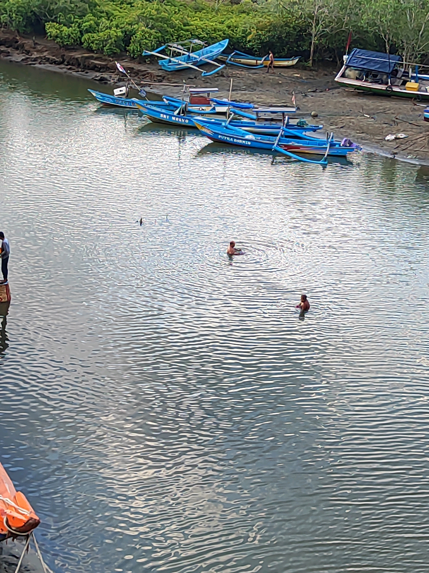 This image depicts the shore of a body of water, likely a river or bay, in Prigi, Indonesia. The scene is populated with several blue fishing boats docked along the muddy bank, which is overgrown with lush green vegetation. The water itself is calm with rippling reflections of the sky and surrounding environment. In the water, two individuals are present. One person is partially submerged and appears to be swimming or playing. Another individual is standing in the water, further away. On the left edge of the frame, a person stands on a structure, possibly a small pier or platform, overlooking the water. Another orange life preserver is visible attached to the platform. The overall impression is a serene, everyday scene at a coastal or riverine location.