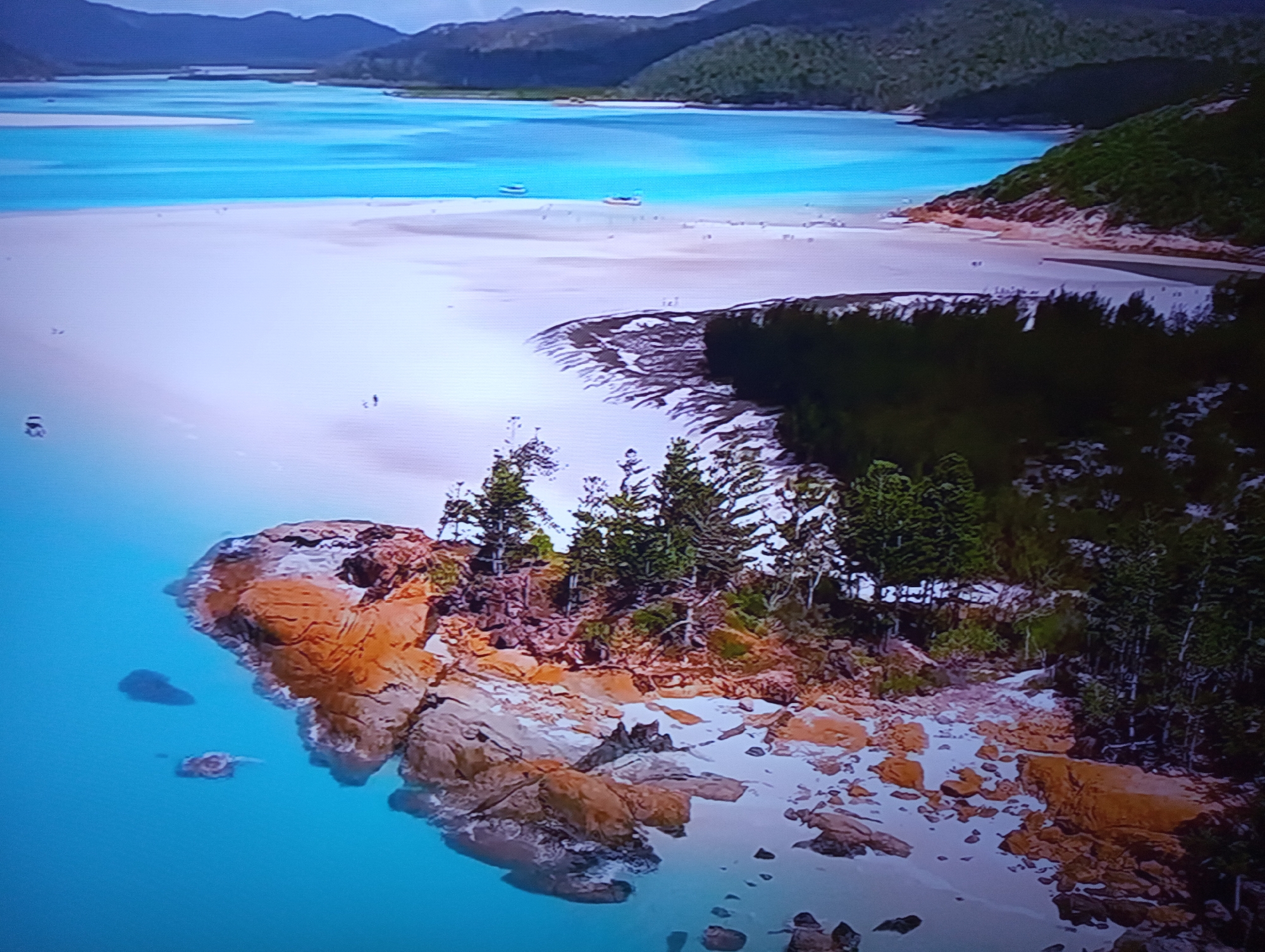 The image depicts a breathtaking coastal landscape with vibrant turquoise waters, a vast expanse of white sand, and lush green hills in the background. The scene is an aerial view of a tropical island, possibly a well-known tourist destination. The foreground features a rocky outcrop with sparse vegetation, jutting into the clear blue water. Scattered on the sand are a few small figures, suggesting the presence of people. In the distance, several boats can be seen on the water, and the horizon is dominated by rolling hills or mountains. The overall impression is one of pristine natural beauty and tranquility.
The image does not depict any recognizable landmarks or elements associated with the city of Dnipro, Ukraine. The landscape is distinctly tropical and coastal, which is not characteristic of Dnipro's geography.